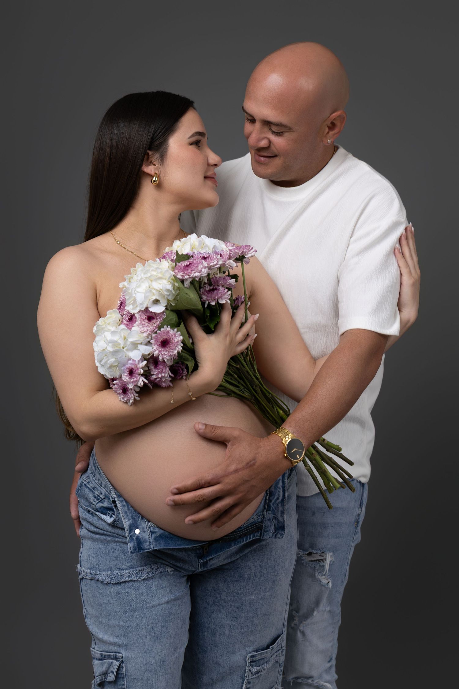 Pregnant couple posing together, woman holding pink and white flowers bouquet, man tenderly touching her baby bump, gray background.