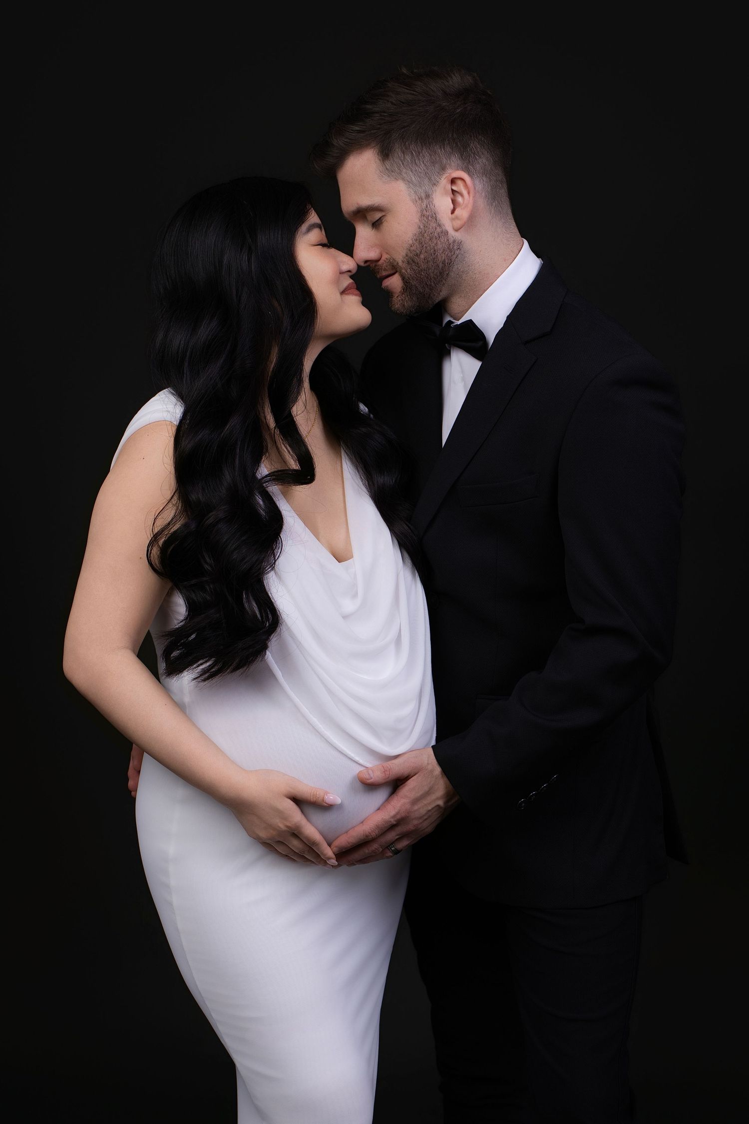 Elegant pregnant couple in formal wear, man in tuxedo embracing woman in white dress against black background.