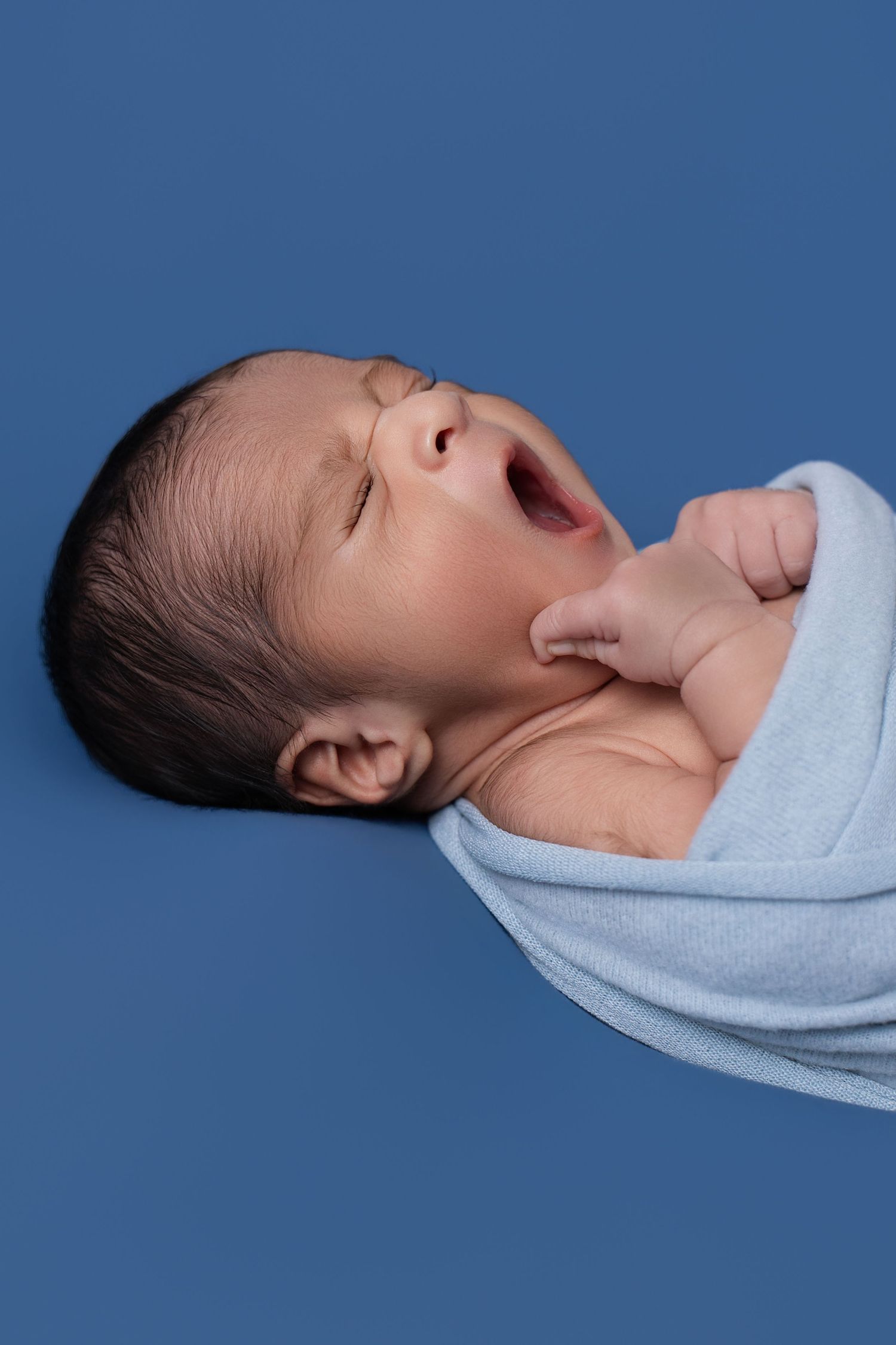 Newborn baby wrapped in light blue swaddle yawning with eyes closed against a blue background.