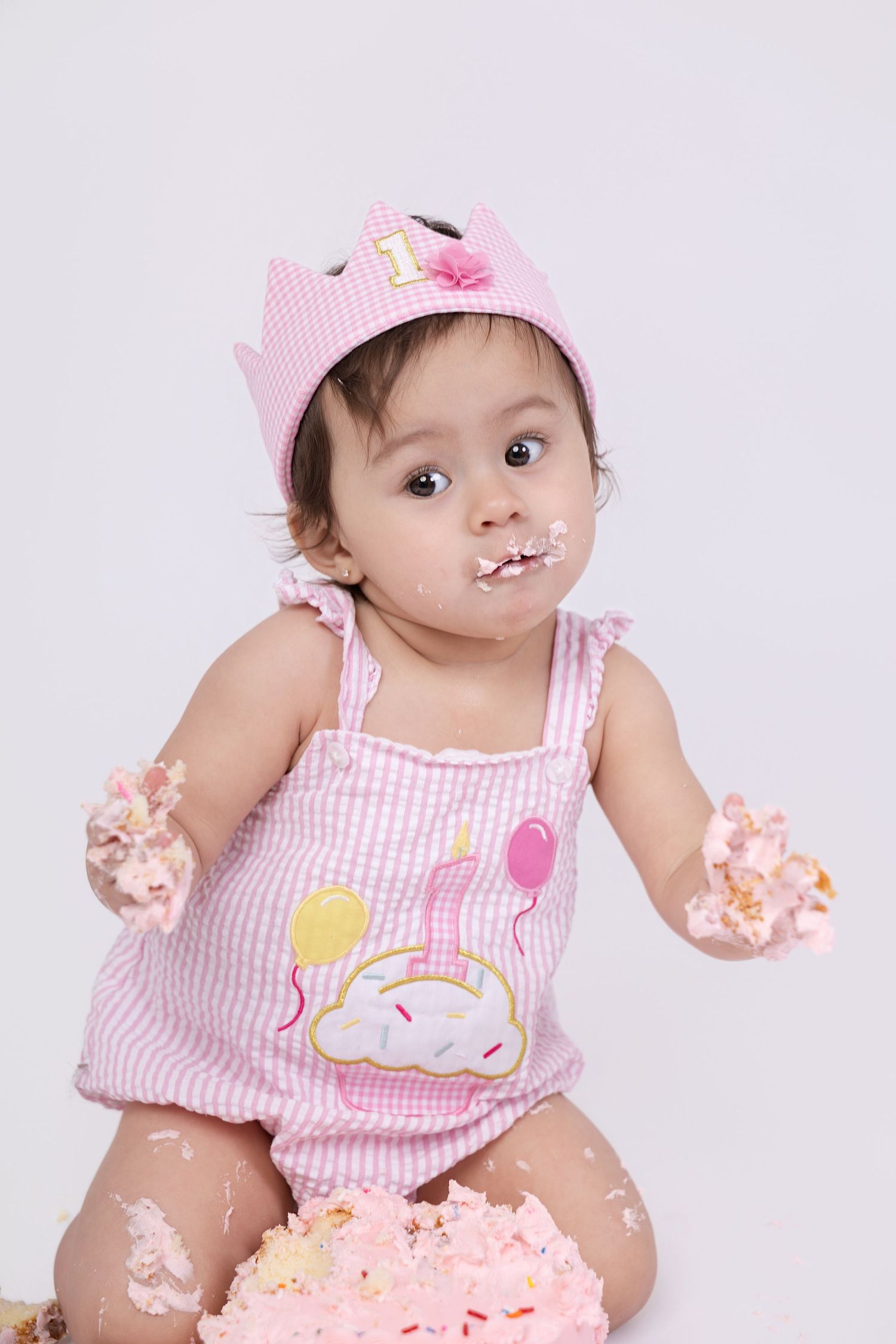 Baby girl in pink crown and dress smashes her first birthday cake, covered in frosting.