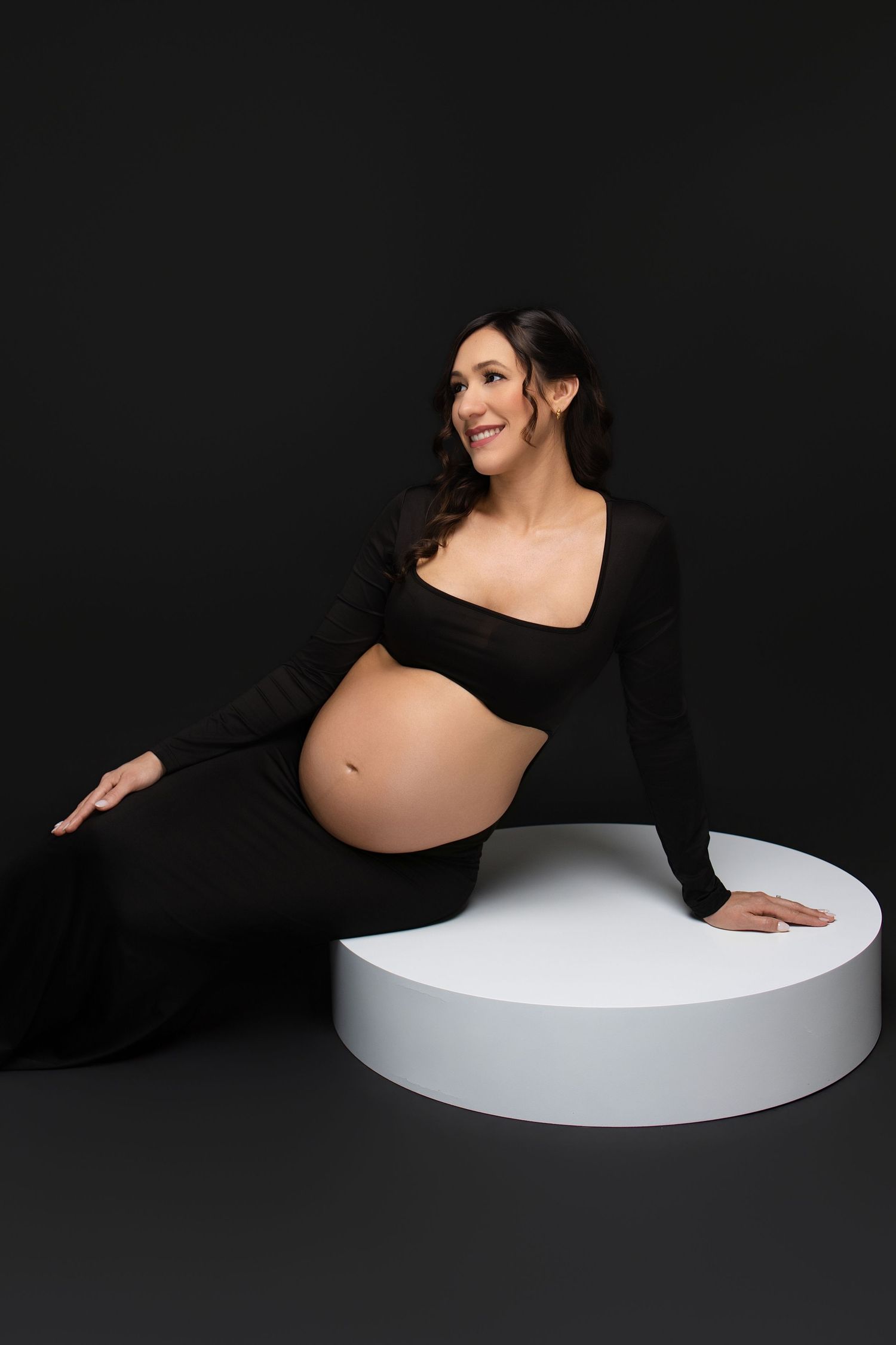 Smiling pregnant woman in black crop top and skirt poses elegantly against a dark studio backdrop.