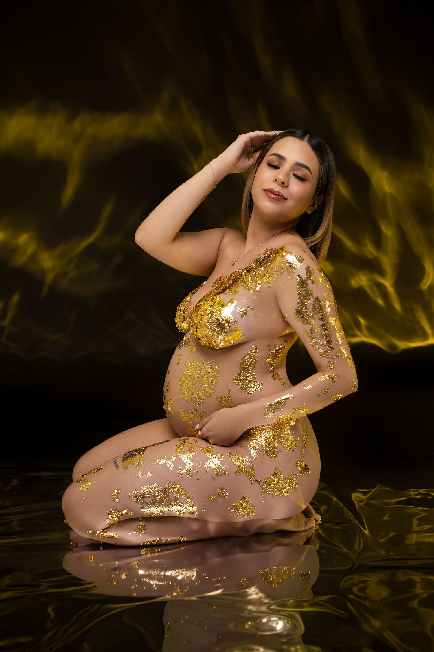 Pregnant woman adorned with gold leaf body paint poses gracefully on reflective surface against dramatic dark golden background.