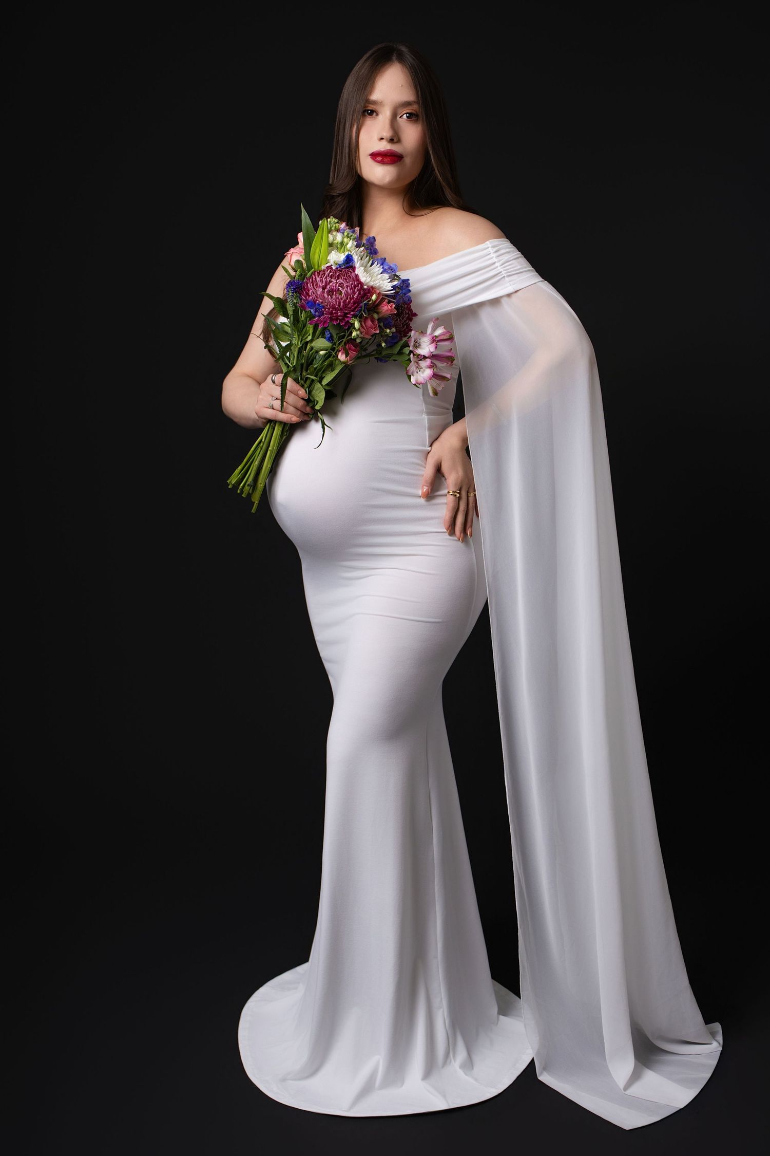Elegant maternity portrait series in white flowing gown against black backdrop holding vibrant floral bouquet.