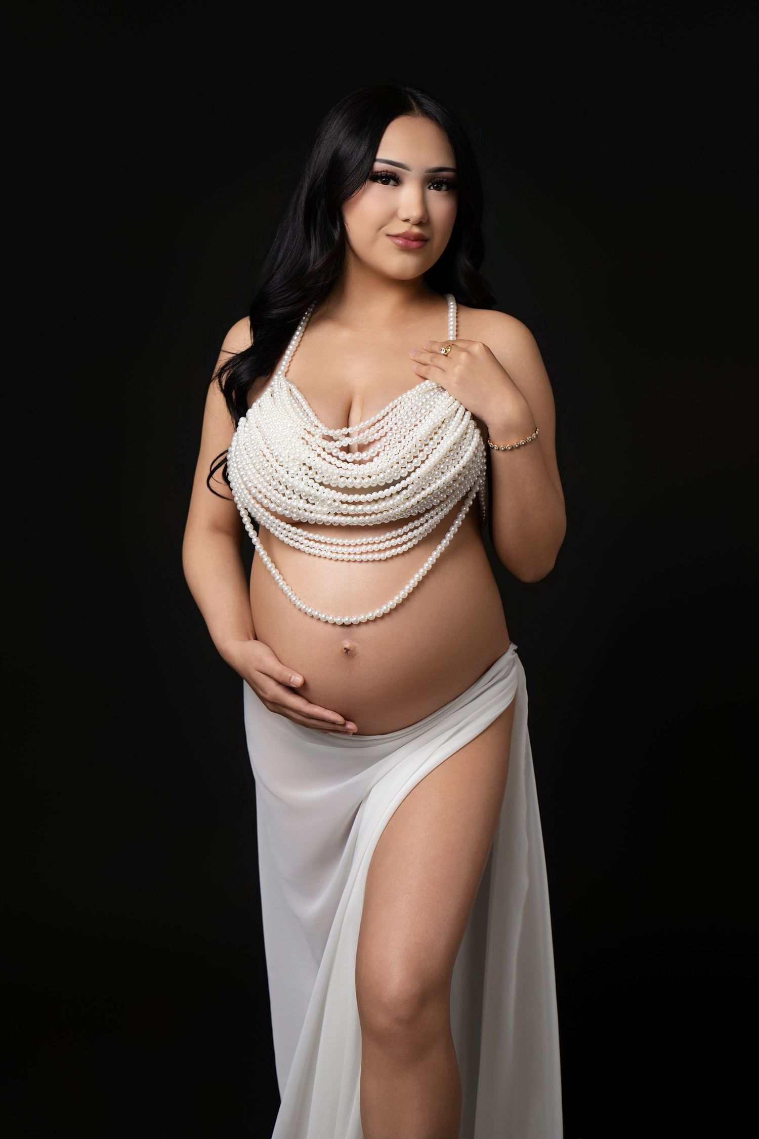 Pregnant woman in pearl top and white skirt posing elegantly against black background in maternity photo shoot.