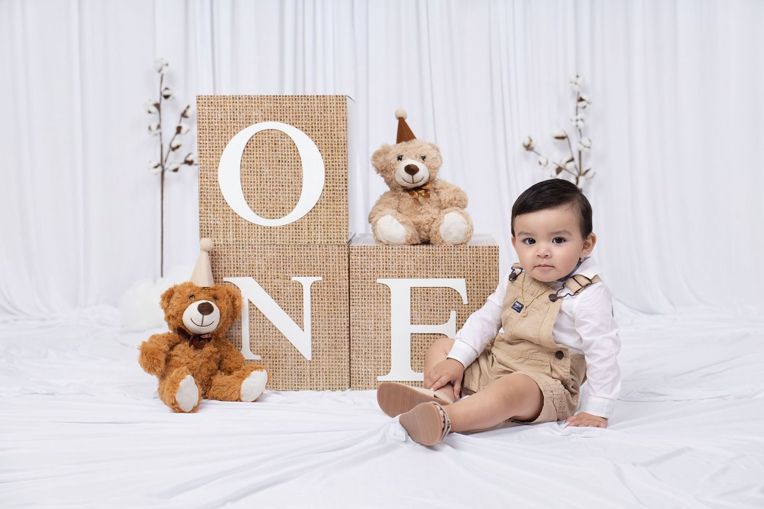Baby boy in tan overalls sits beside burlap ONE blocks with teddy bears for a first birthday photo shoot.