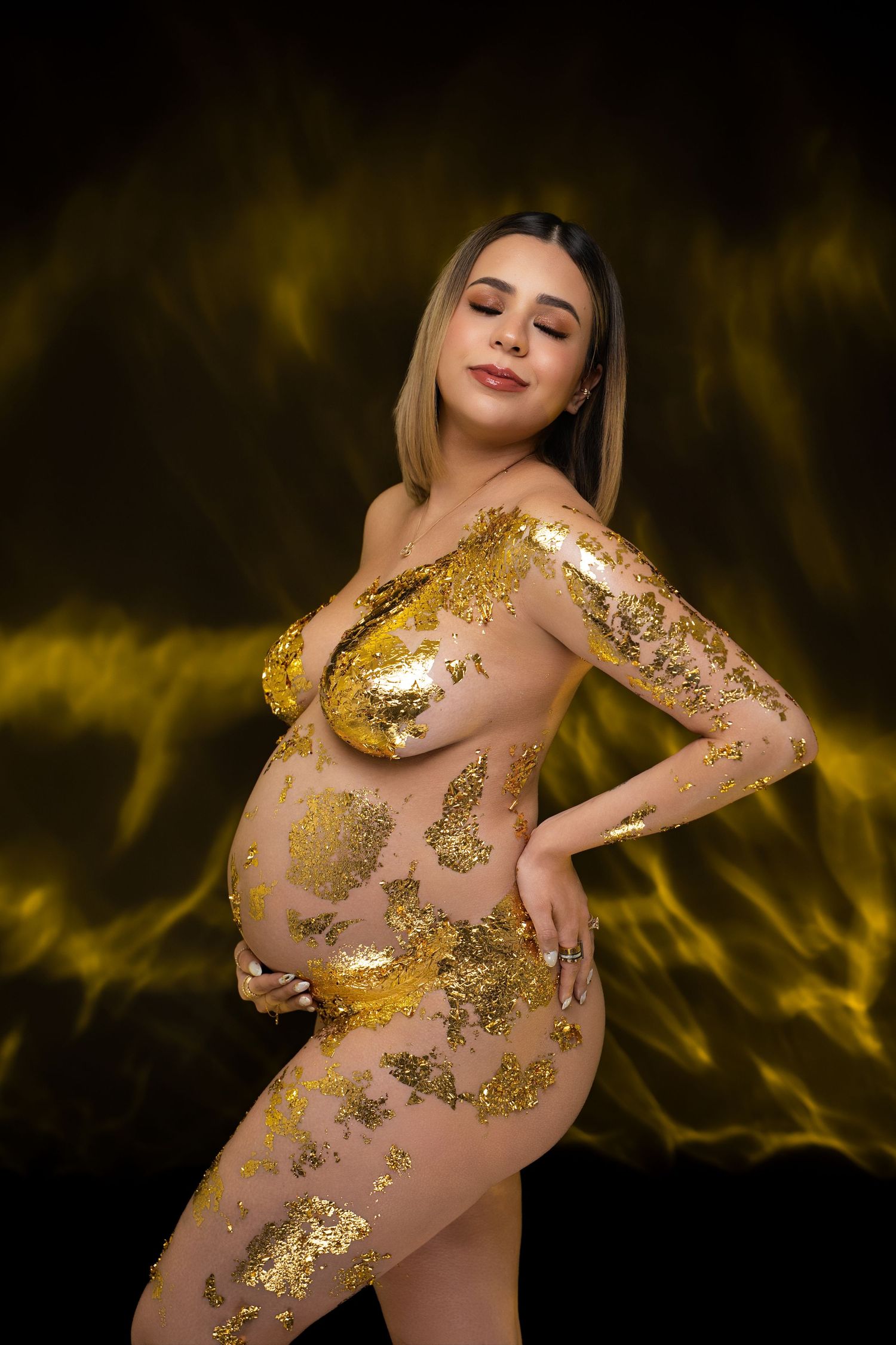 Pregnant woman poses with gold foil body art against dramatic dark golden background in artistic maternity photo.