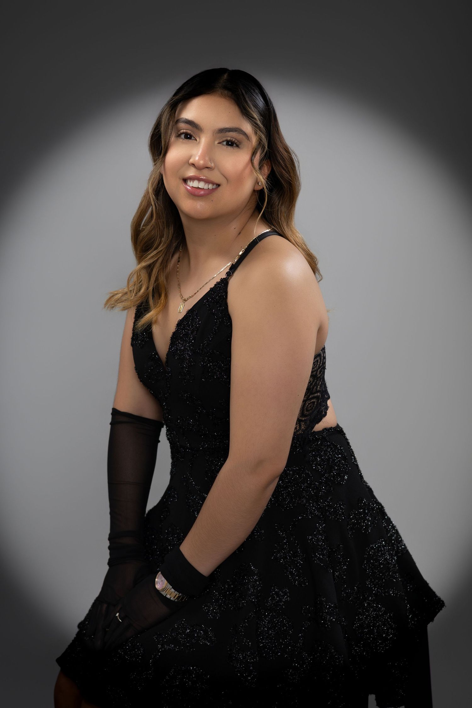 Young woman in sparkling black gown and gloves poses elegantly against gray studio backdrop.