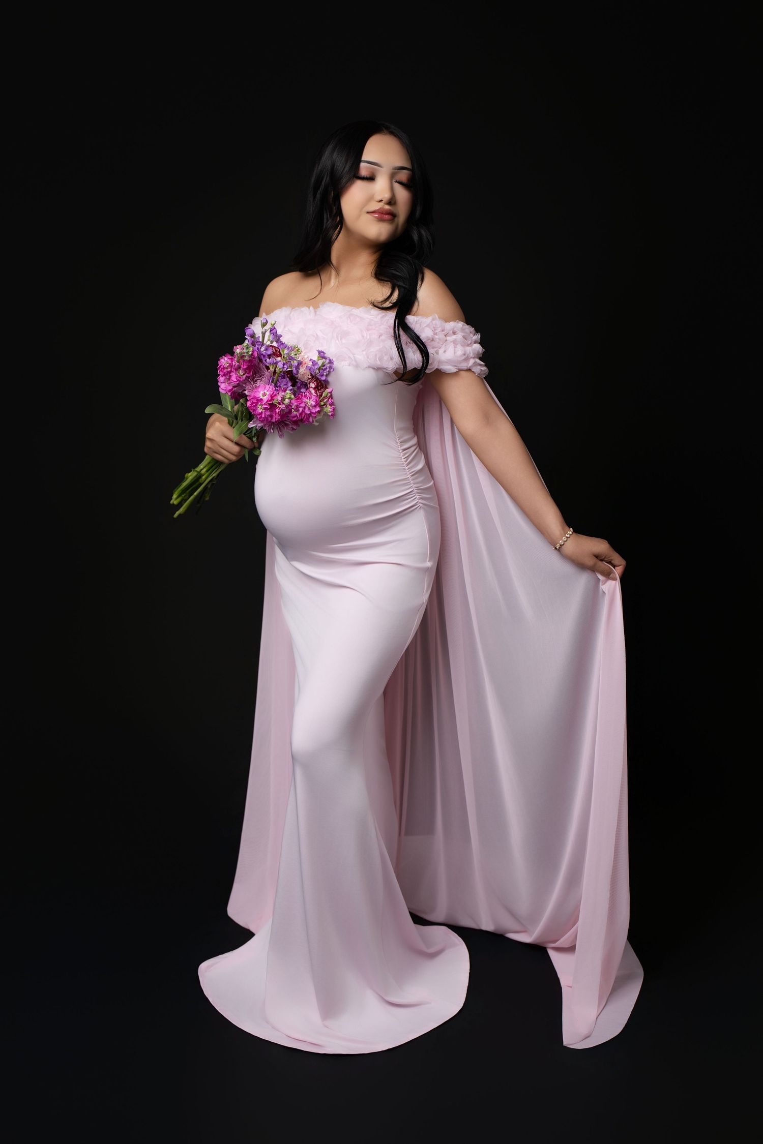Pregnant woman in pink off-shoulder gown holding purple floral bouquet, posing against black background.
