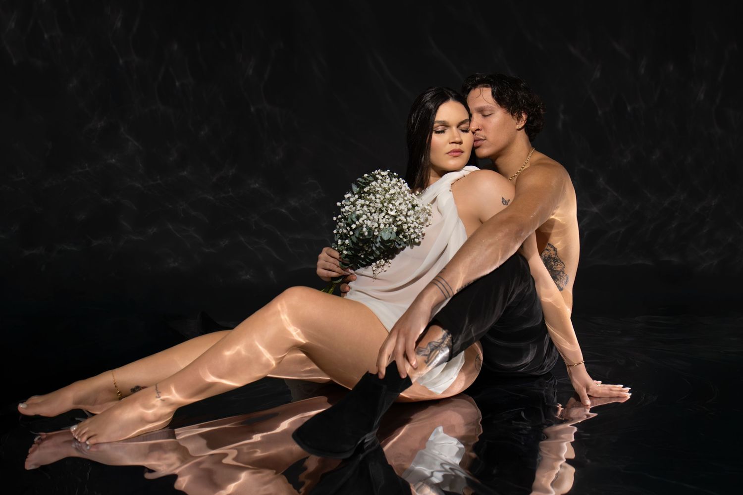 A couple poses intimately on a reflective floor, woman holding white flowers, against a dark studio background.