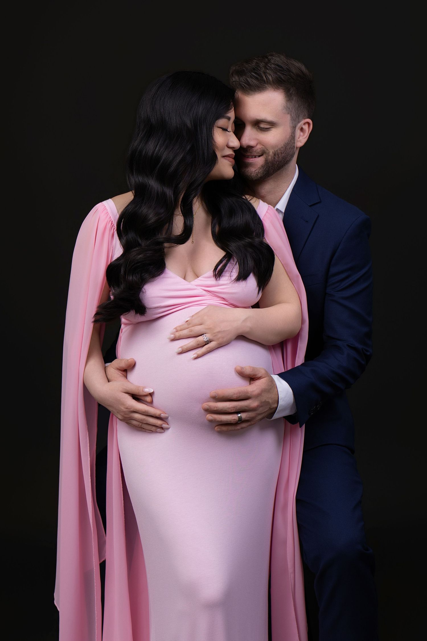 A man in a navy suit embraces a pregnant woman in a pink gown, both smiling tenderly against a dark background.