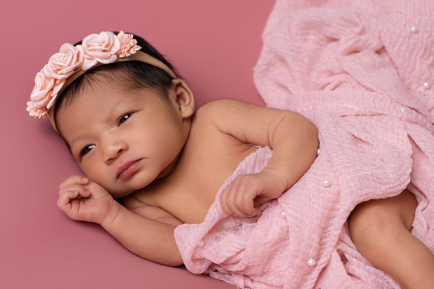 Newborn baby girl wearing a pink floral headband, wrapped in a soft pink blanket on a pink background.