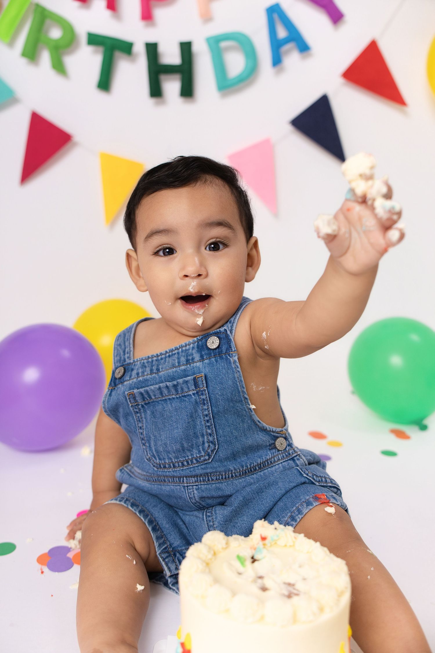 Baby boy in denim overalls smashing first birthday cake, surrounded by colorful balloons and Happy Birthday banner.