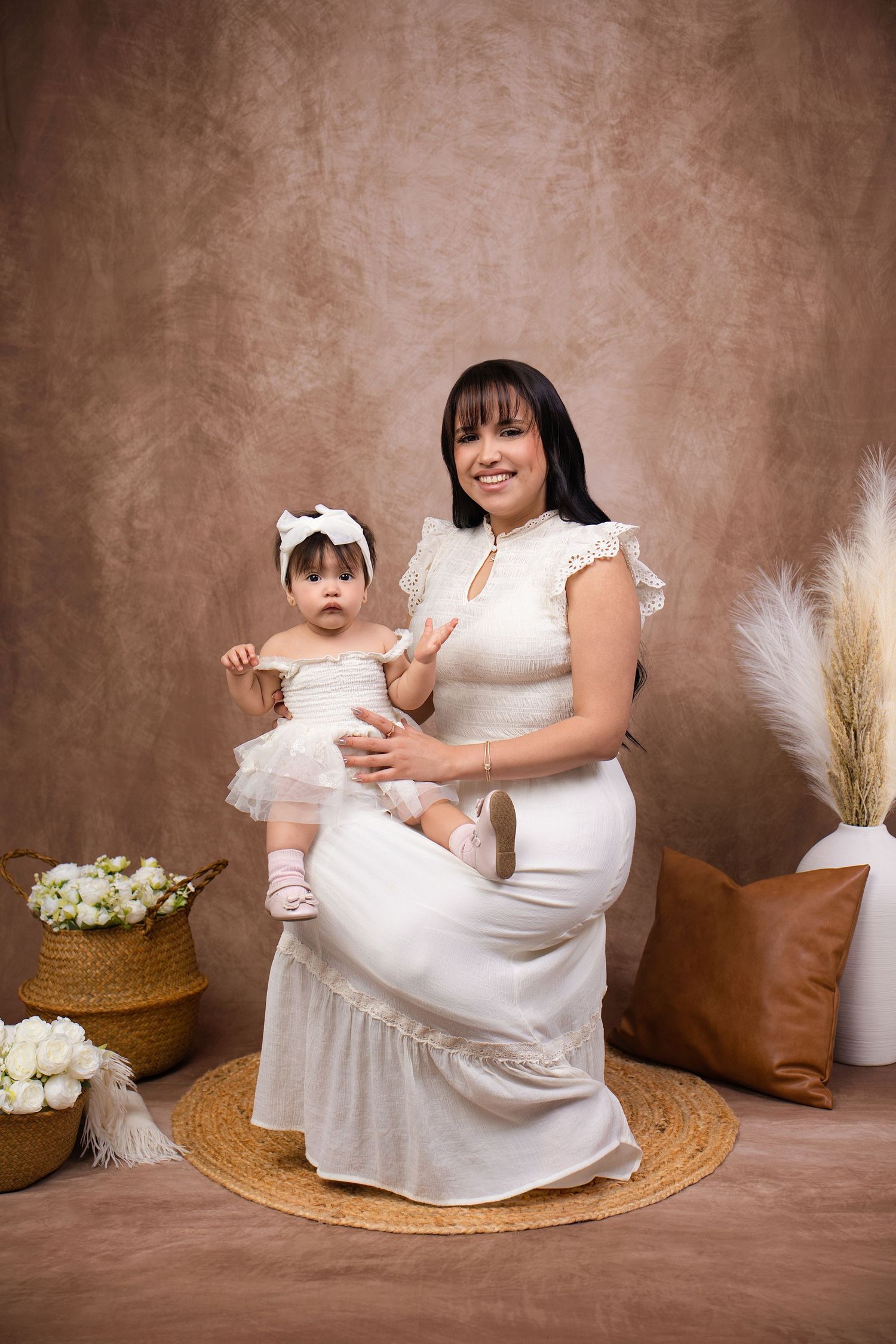 Mother and baby girl in matching white dresses pose together for a studio portrait photo.