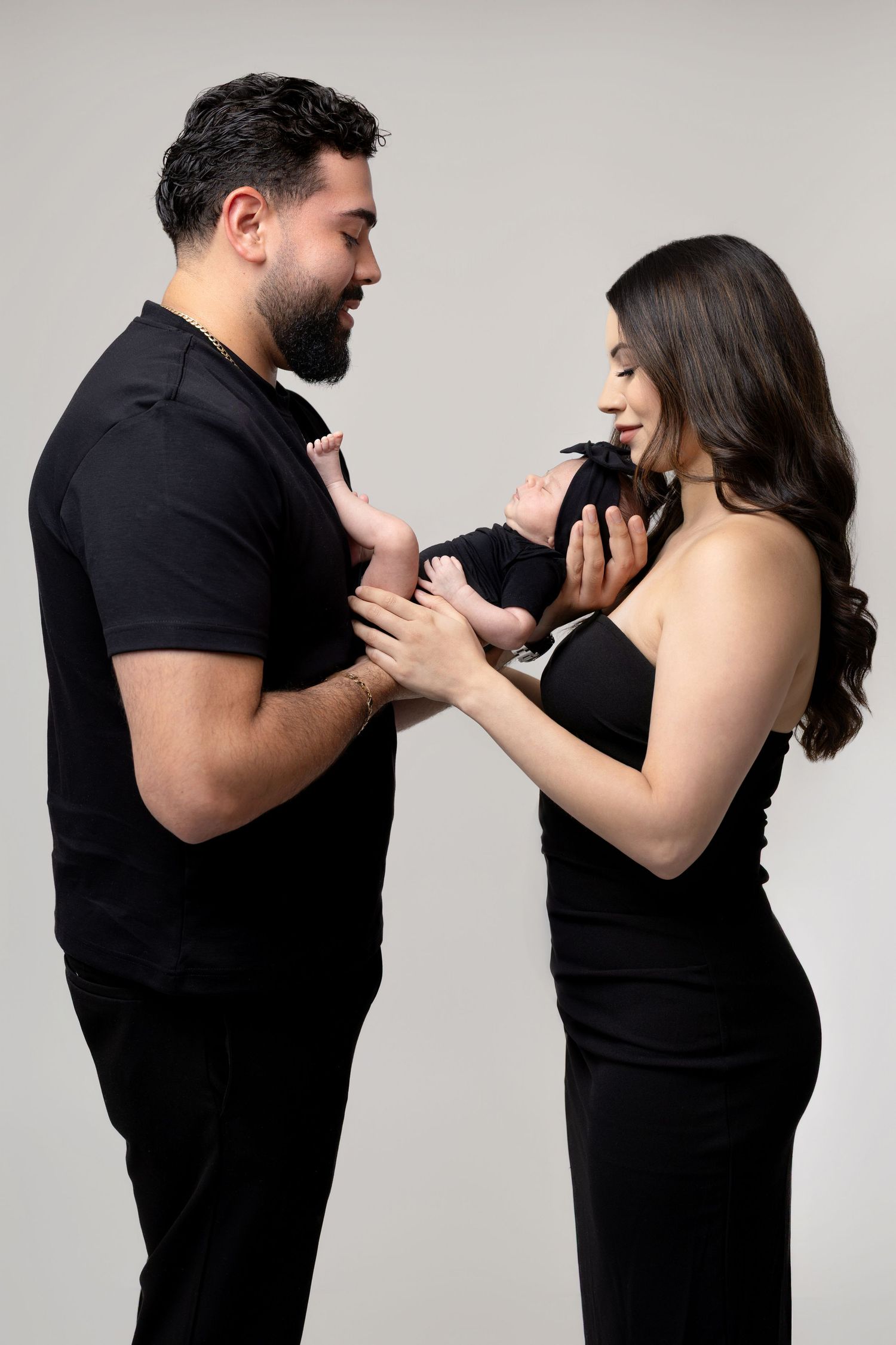 A couple dressed in black holds their newborn baby, smiling lovingly at the infant in a studio portrait.
