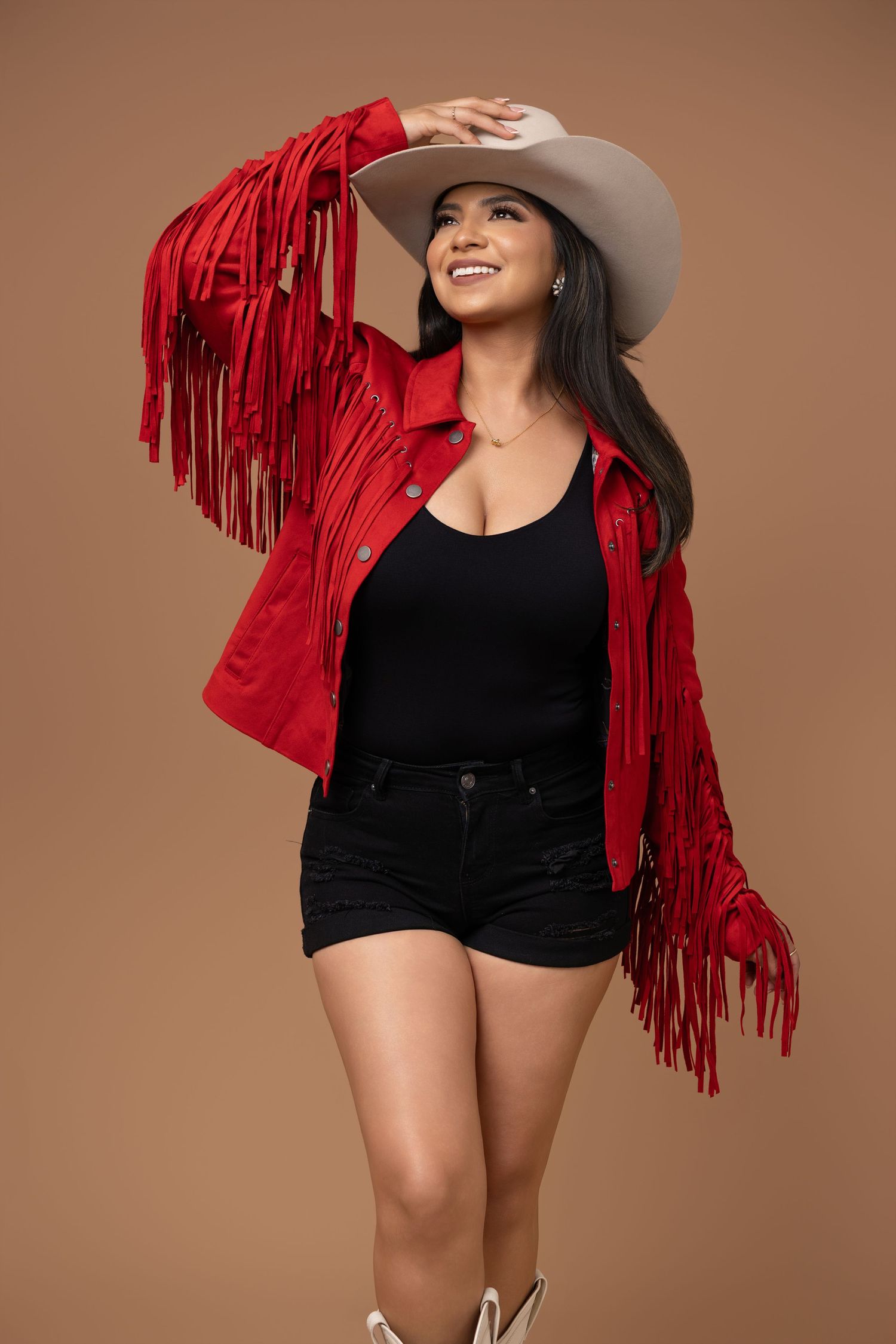 Woman in red fringe jacket, black top, denim shorts, and cowboy hat posing confidently against a tan background.