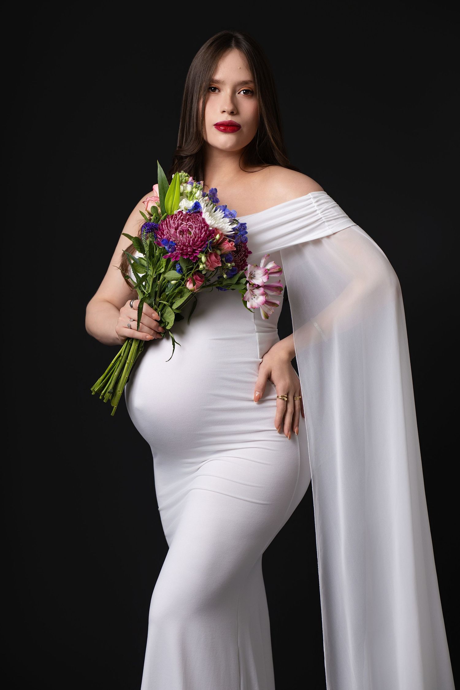 Maternity photo series showing a woman in flowing white dress holding colorful flowers against black background.