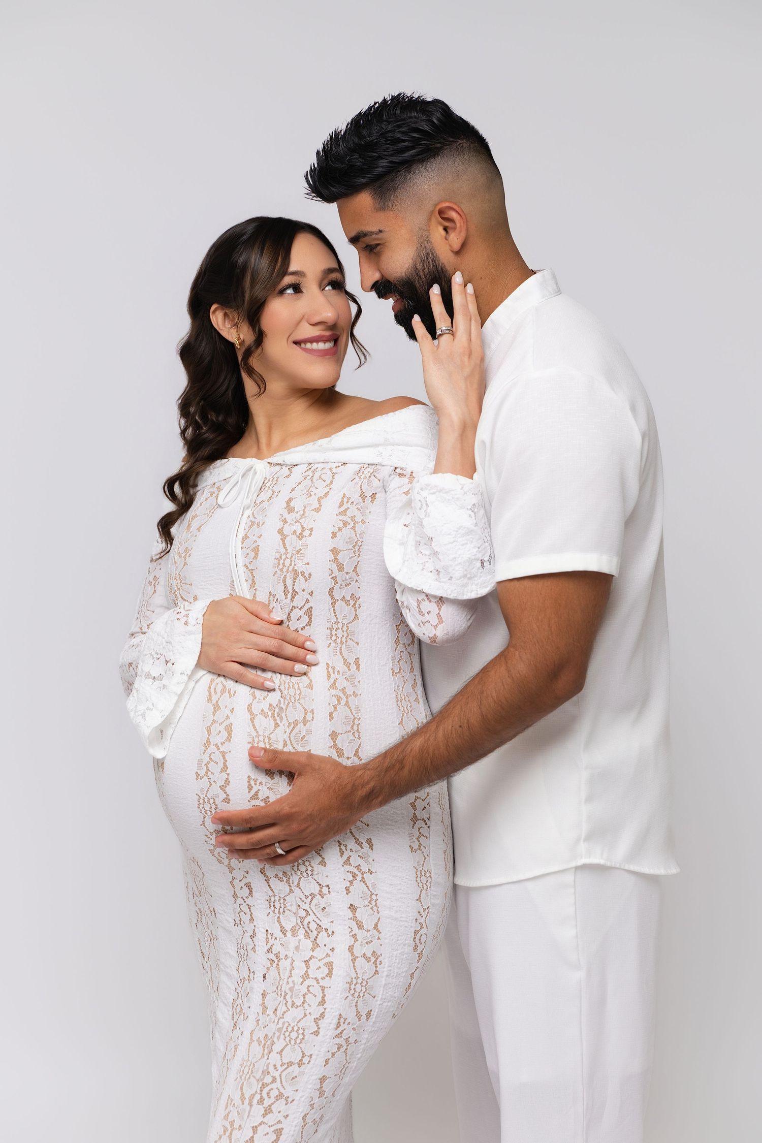 Pregnant couple in white outfits sharing a tender moment, woman in lace dress cradling baby bump.