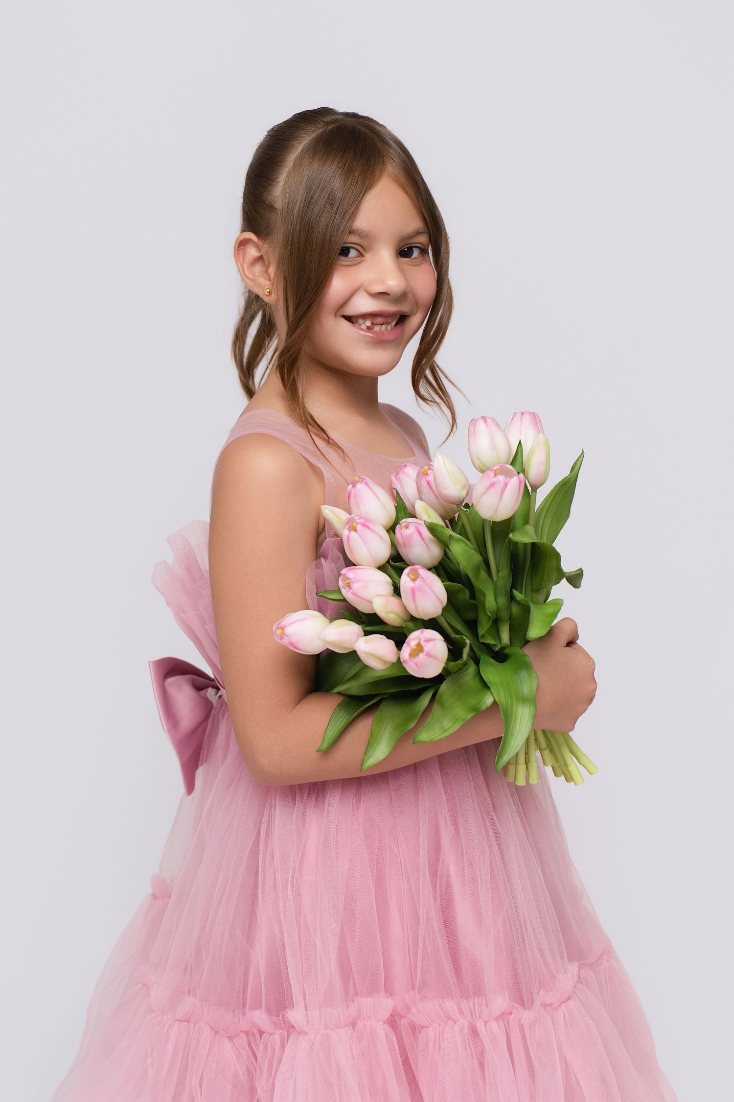 Smiling young girl in pink tulle dress holding bouquet of pink tulips against light gray background.