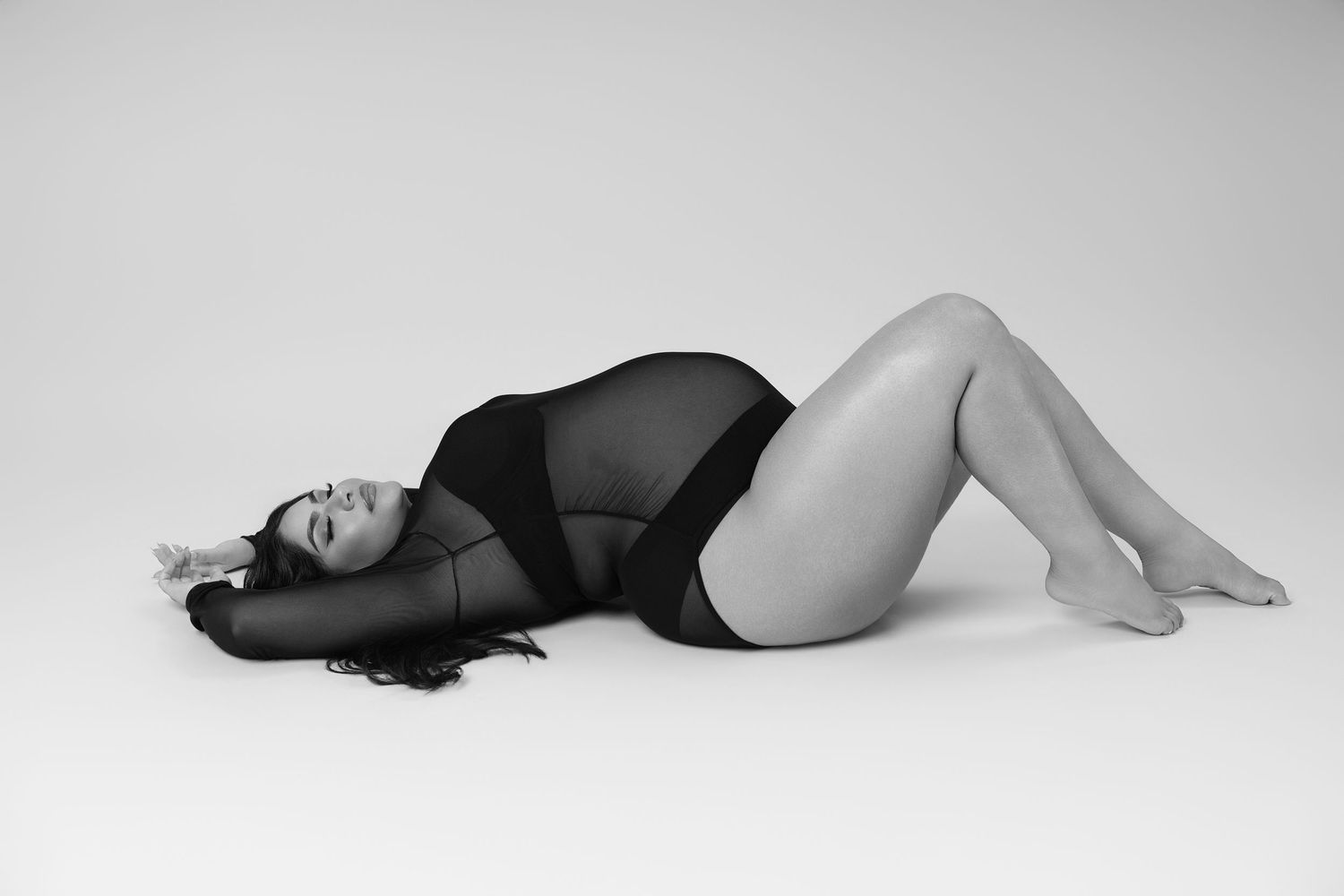 Black and white photo of a curvy woman in a black bodysuit posing gracefully on a studio floor.