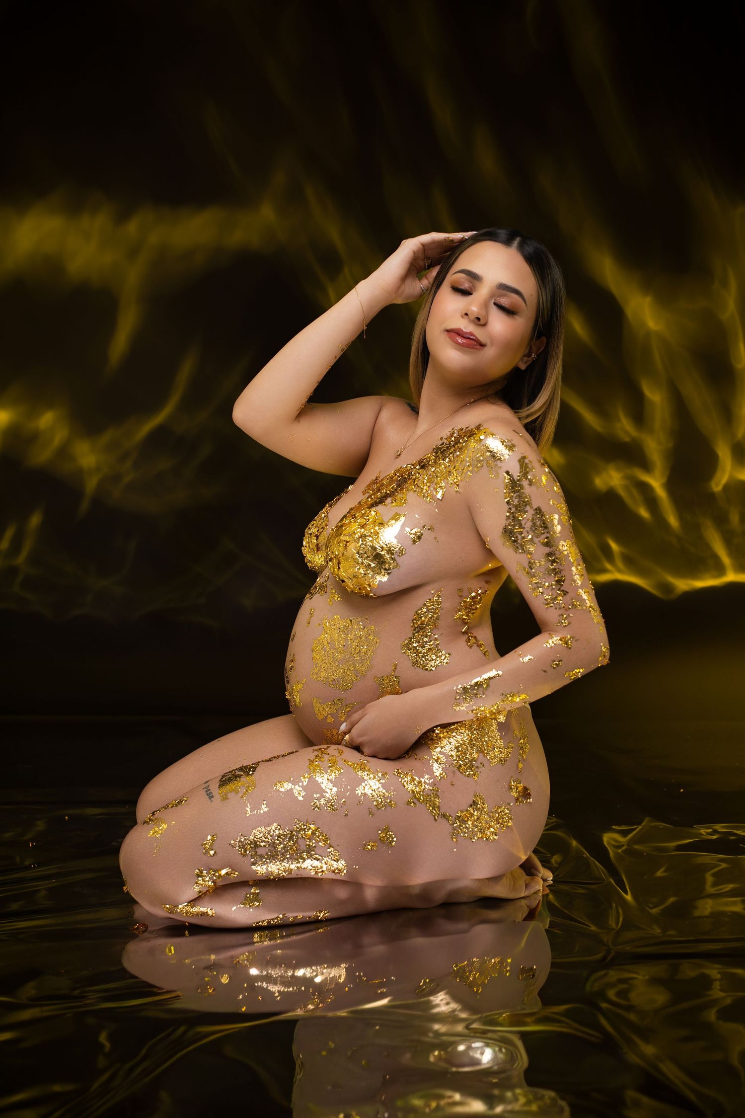 Pregnant woman adorned with gold leaf body art poses gracefully in reflective water against a dramatic dark background.