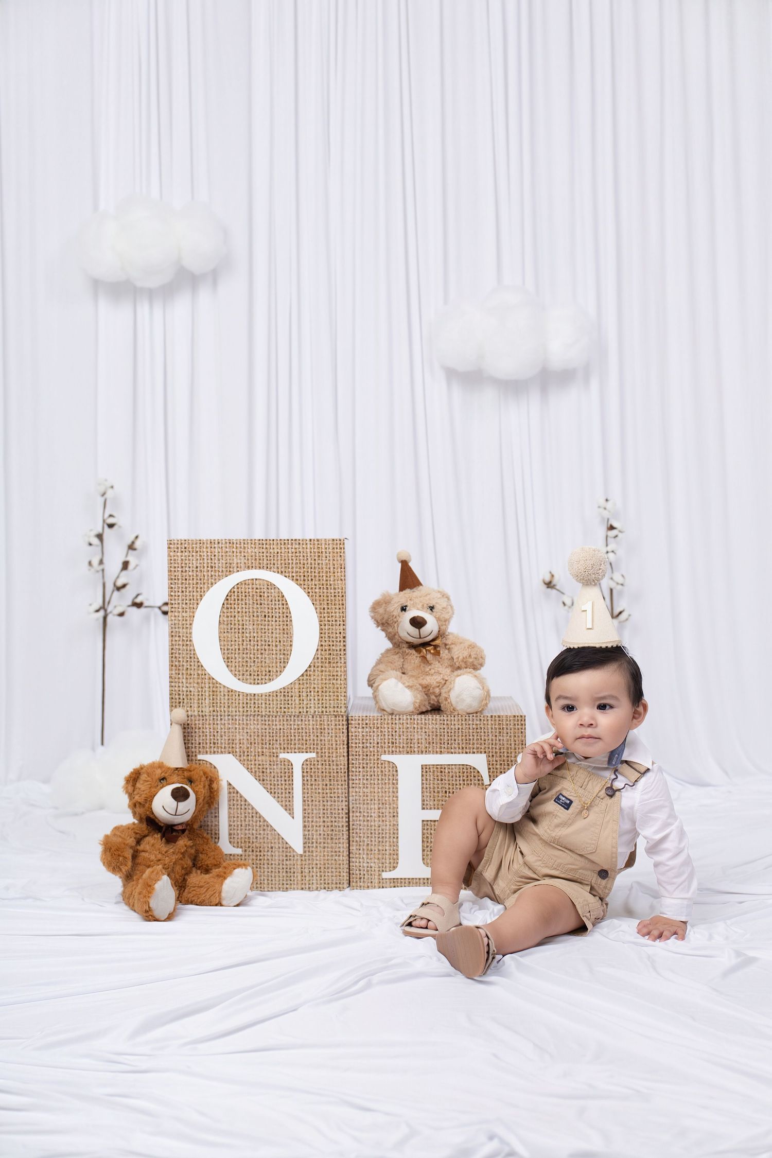 A baby boy sits beside teddy bears and ONE letter blocks at a first birthday photo shoot with white drape backdrop.