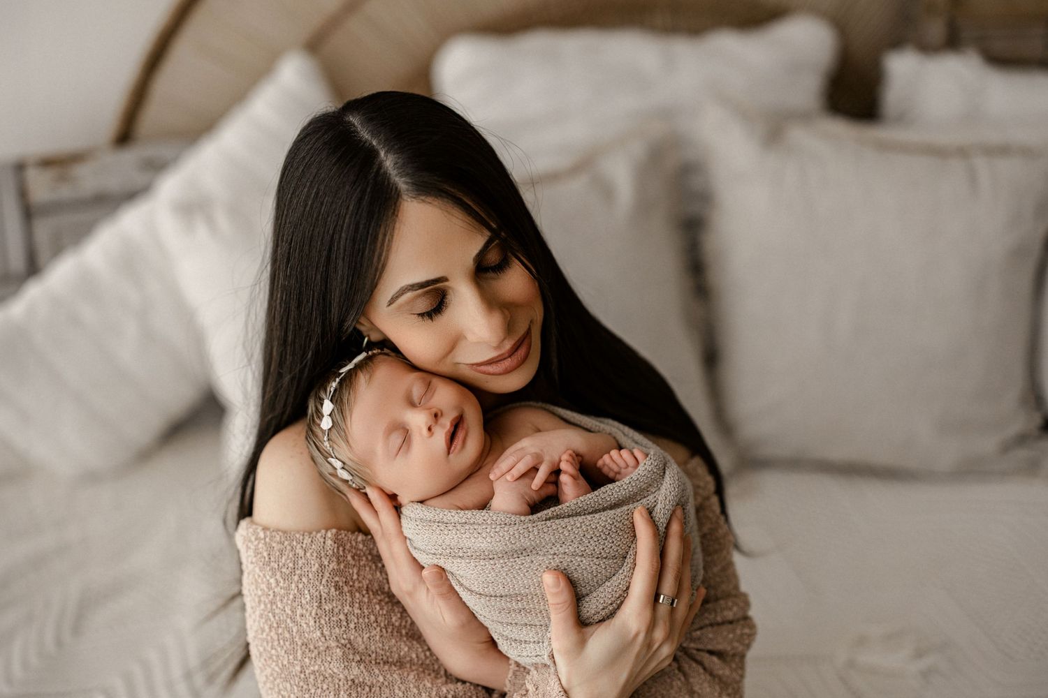 Parent in beige cardigan tenderly holds sleeping swaddled newborn on white bedding in intimate moment.
