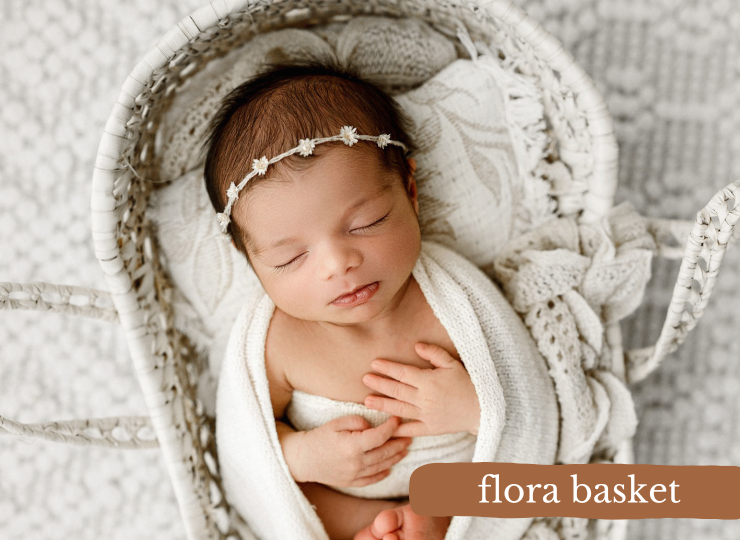 Sleeping newborn baby in a knit flora basket with a floral headband, wrapped in a soft white blanket.