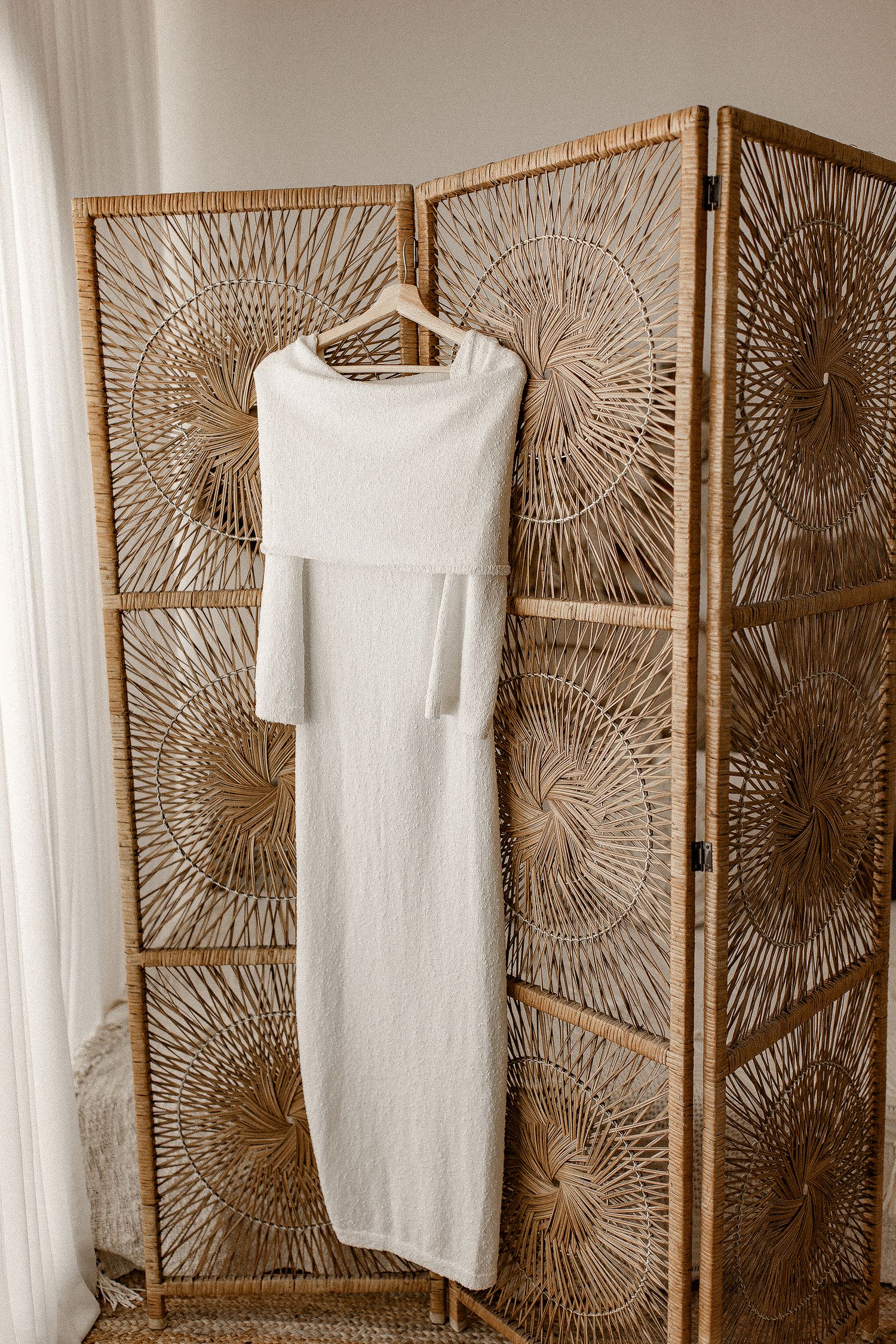 A long white knit dress hangs on a hanger against a decorative rattan room divider with sunburst pattern designs.