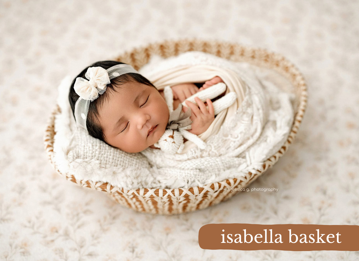 Newborn baby sleeping peacefully in an Isabella wicker basket, wrapped in cream knit blanket with white bow headband.