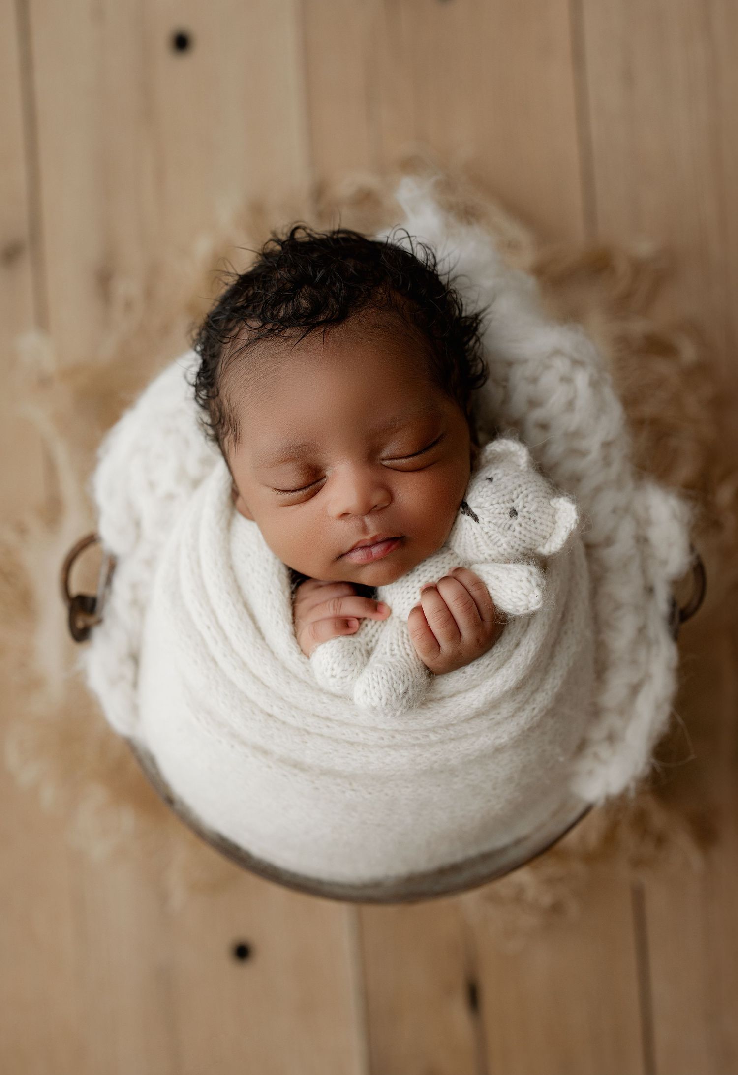 Sleeping newborn wrapped in soft white swaddle blanket with small plush toy in rustic wooden basket.
