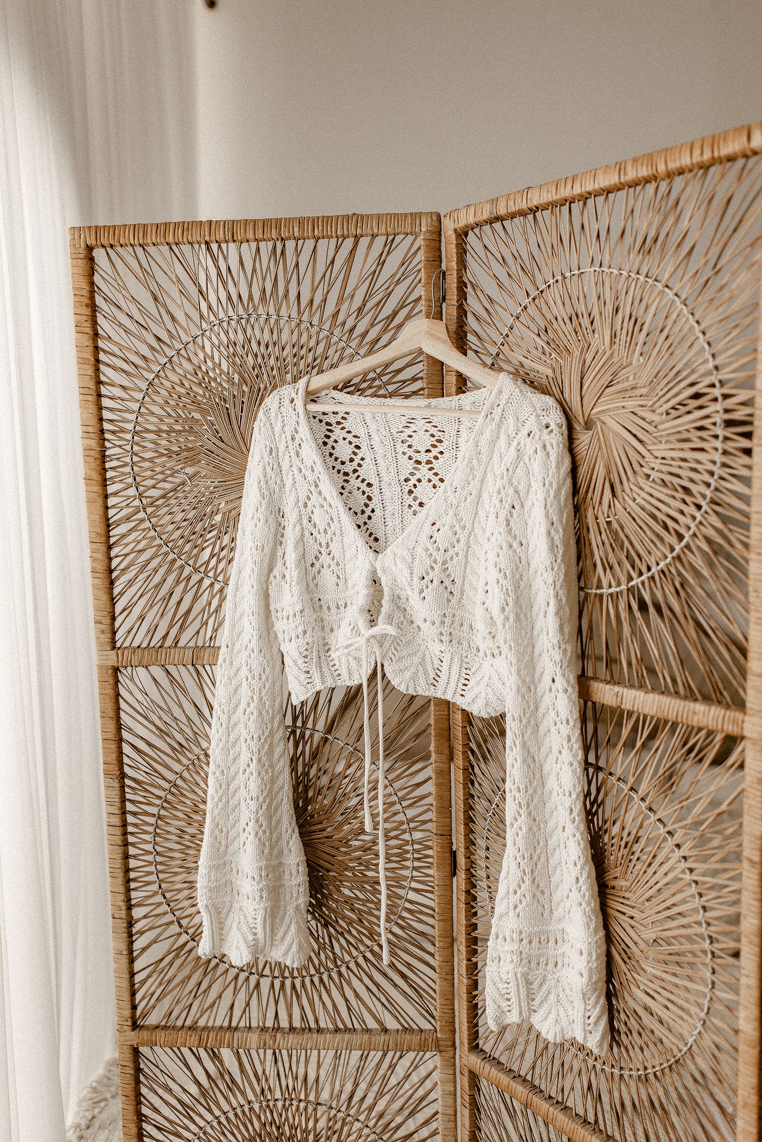 Delicate white crochet crop top with bell sleeves hangs against a woven rattan folding screen with sunburst pattern.