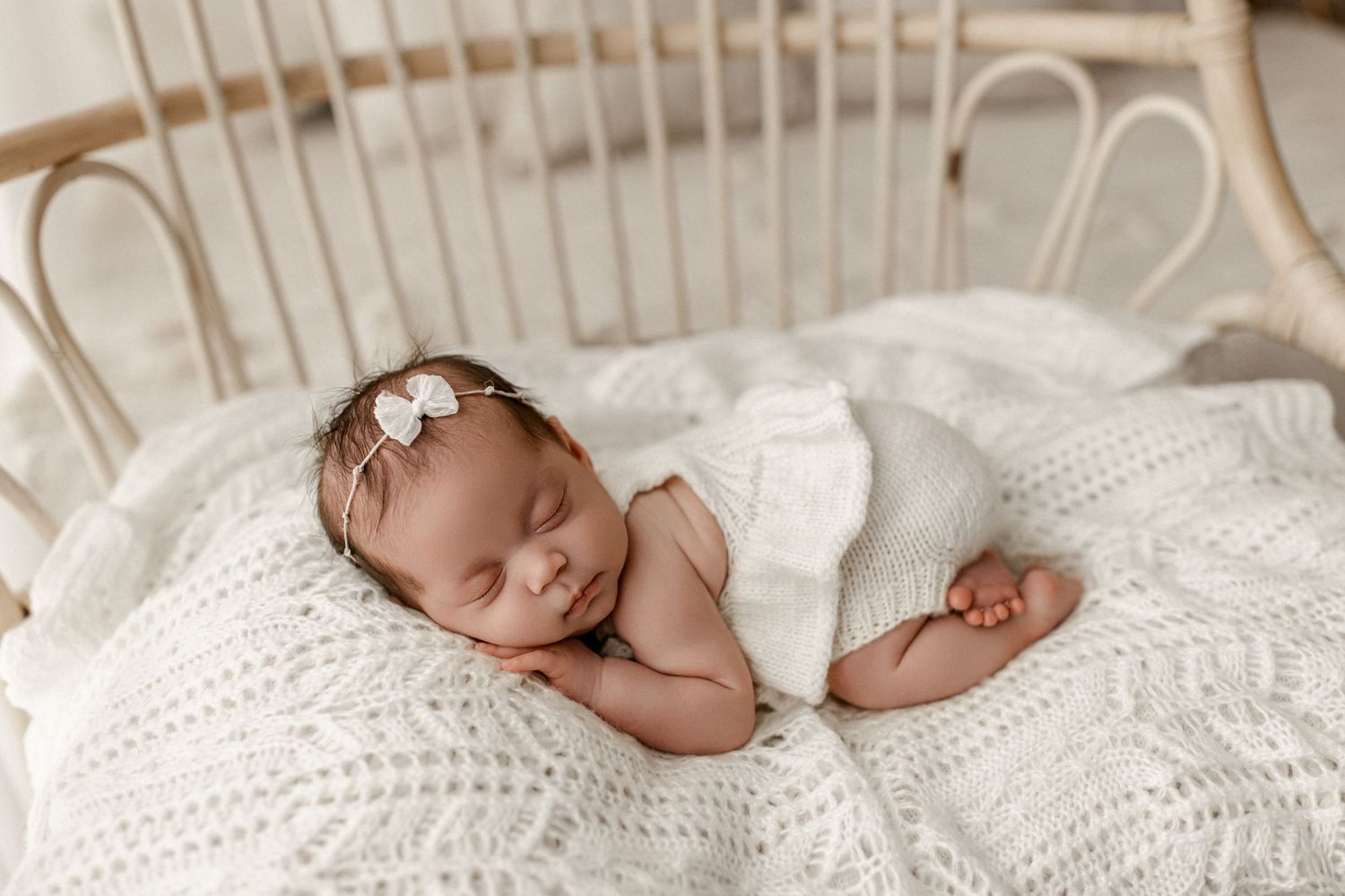 Newborn baby sleeping peacefully on white knitted blanket wearing delicate pearl headband.