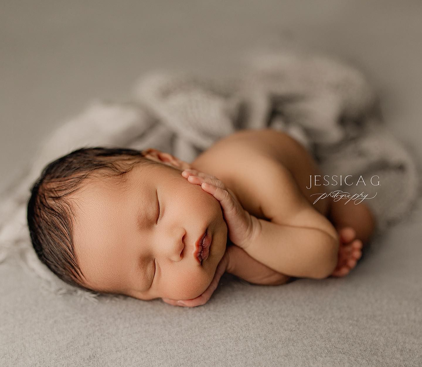 A peaceful sleeping newborn baby lies curled up on soft gray fabric in a professional portrait setting.