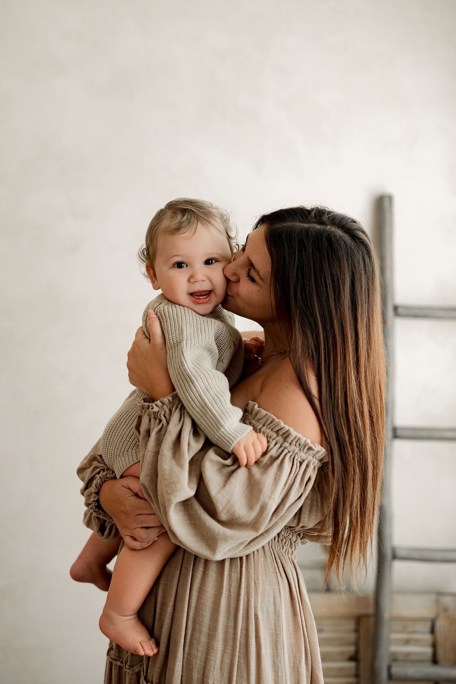 A young mother in a beige off-shoulder dress kisses her smiling baby, both dressed in matching neutral tones.