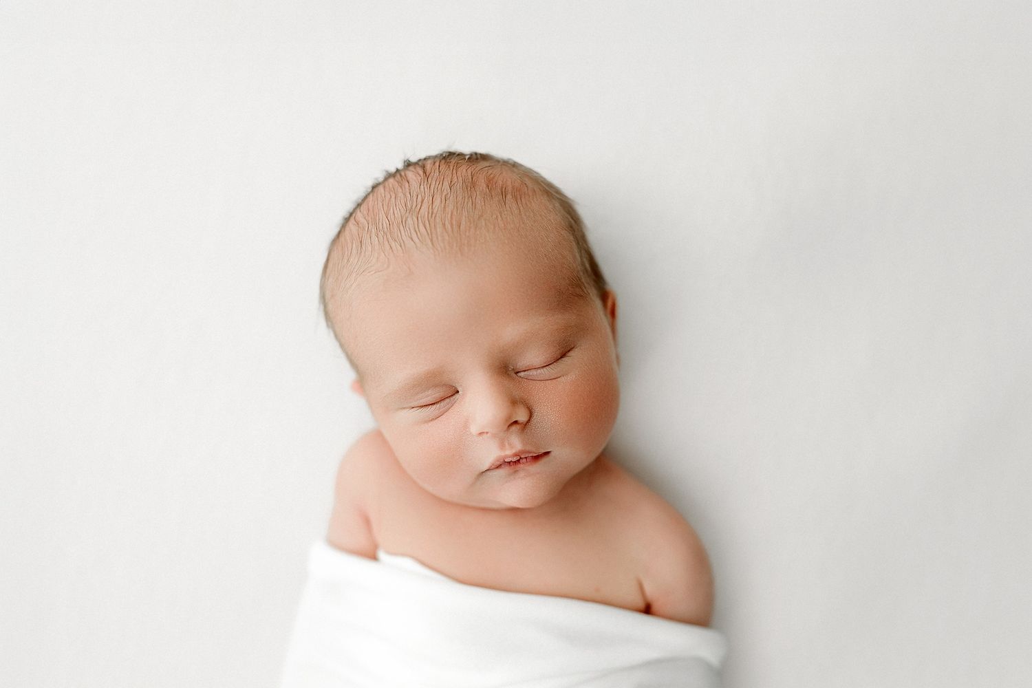 Peaceful sleeping newborn baby wrapped in white fabric against a minimalist white background.