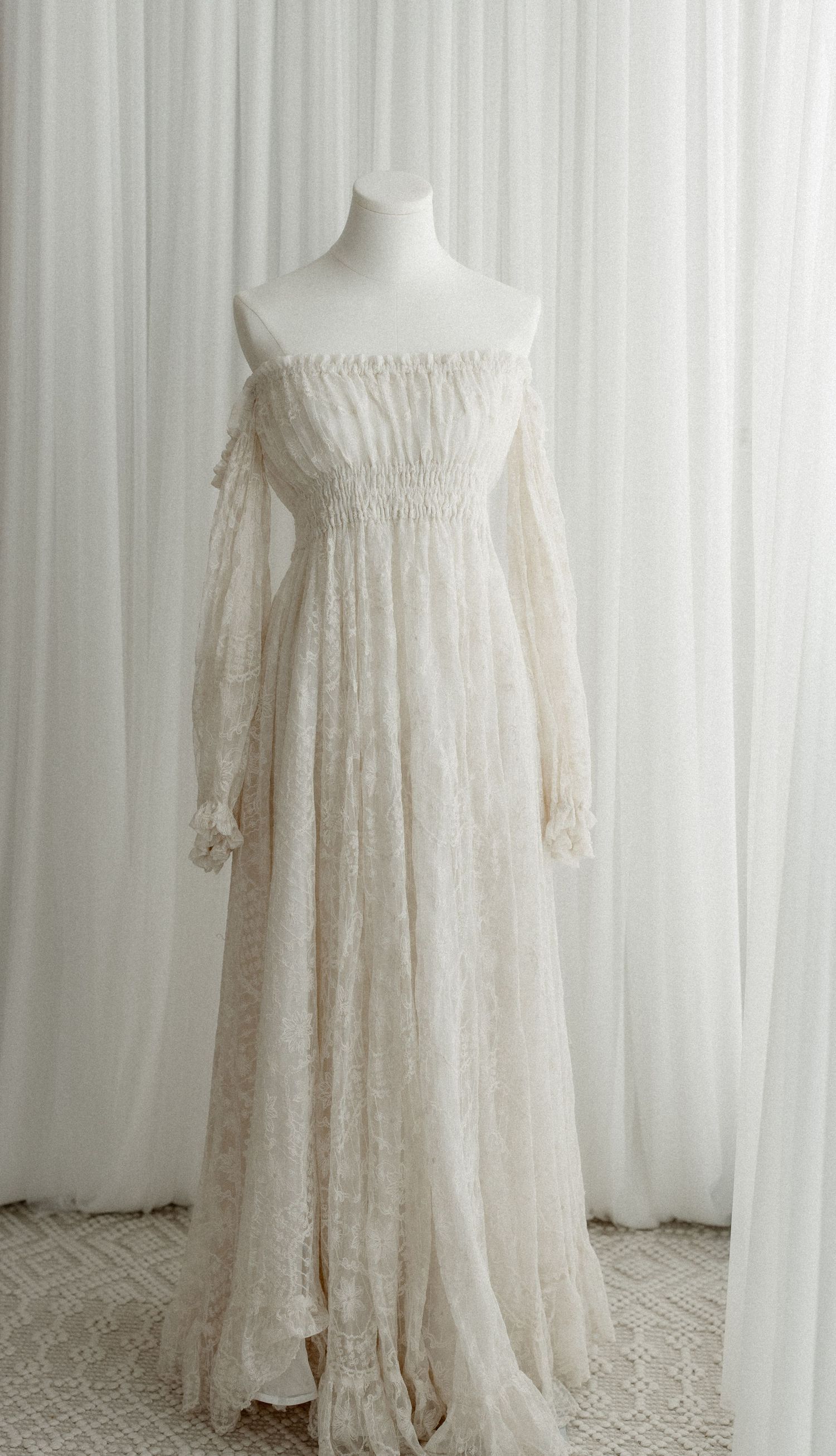 Ethereal off-white lace maternity gown with flowing sleeves and empire waist displayed on mannequin against white backdrop.