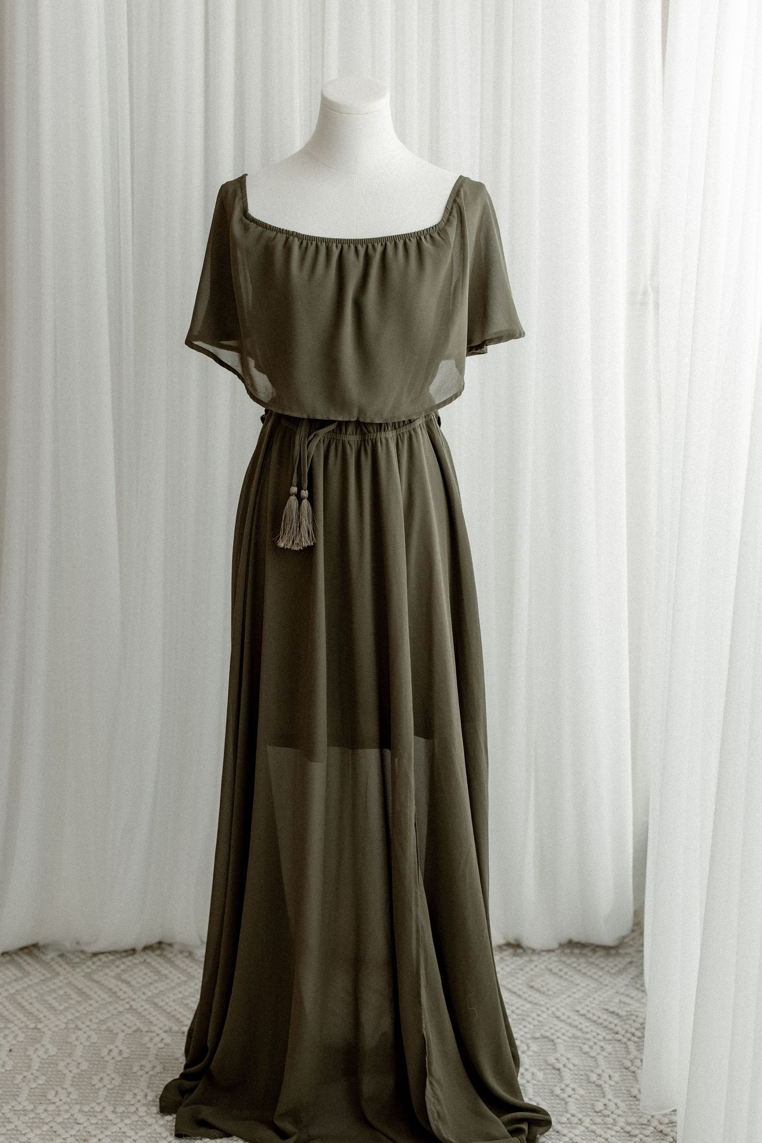 Olive green flowy maxi dress with empire waist and short sleeves displayed on white mannequins against light backdrop.