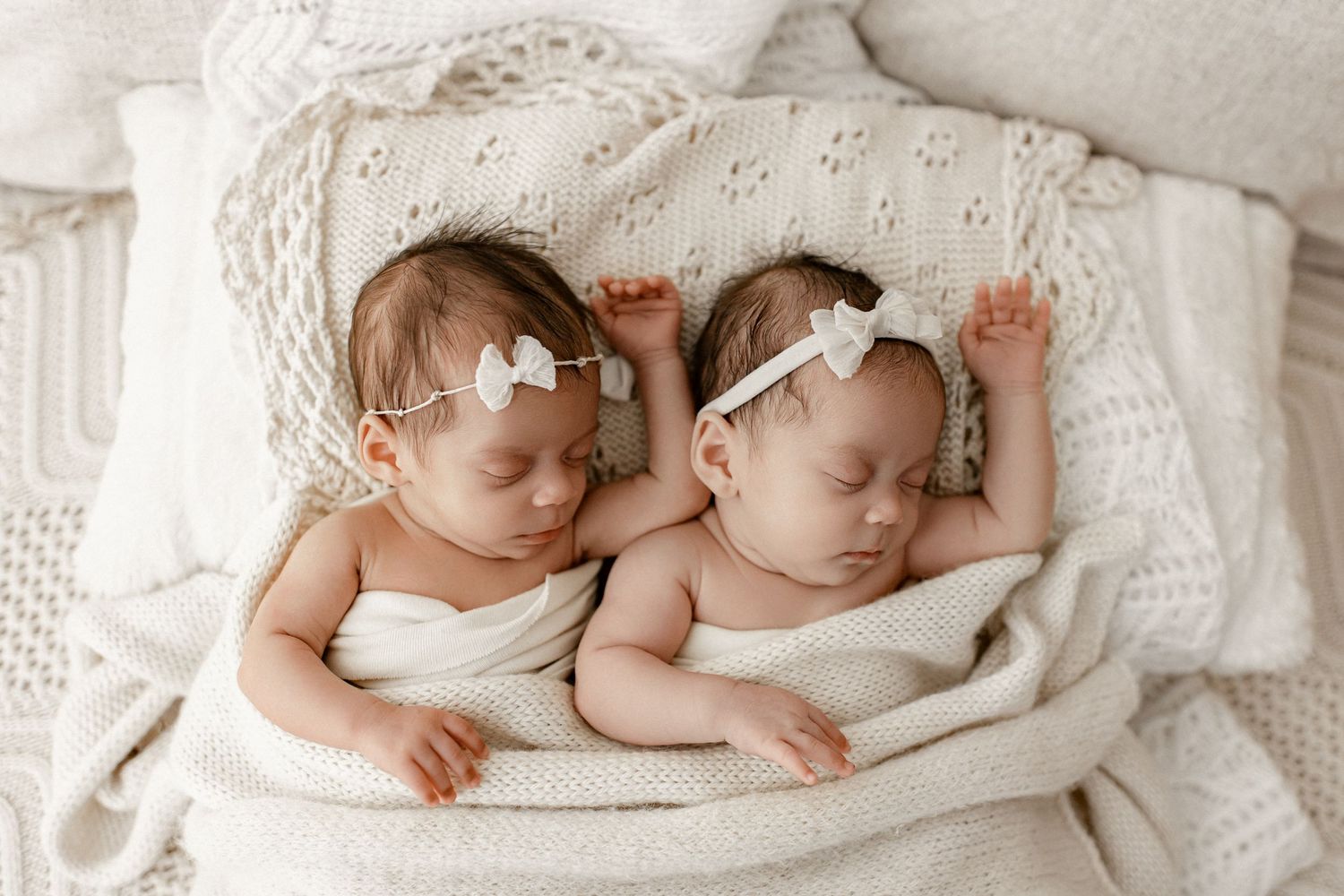 Two sleeping babies wearing white headbands lie side by side on white bedding in a peaceful pose.