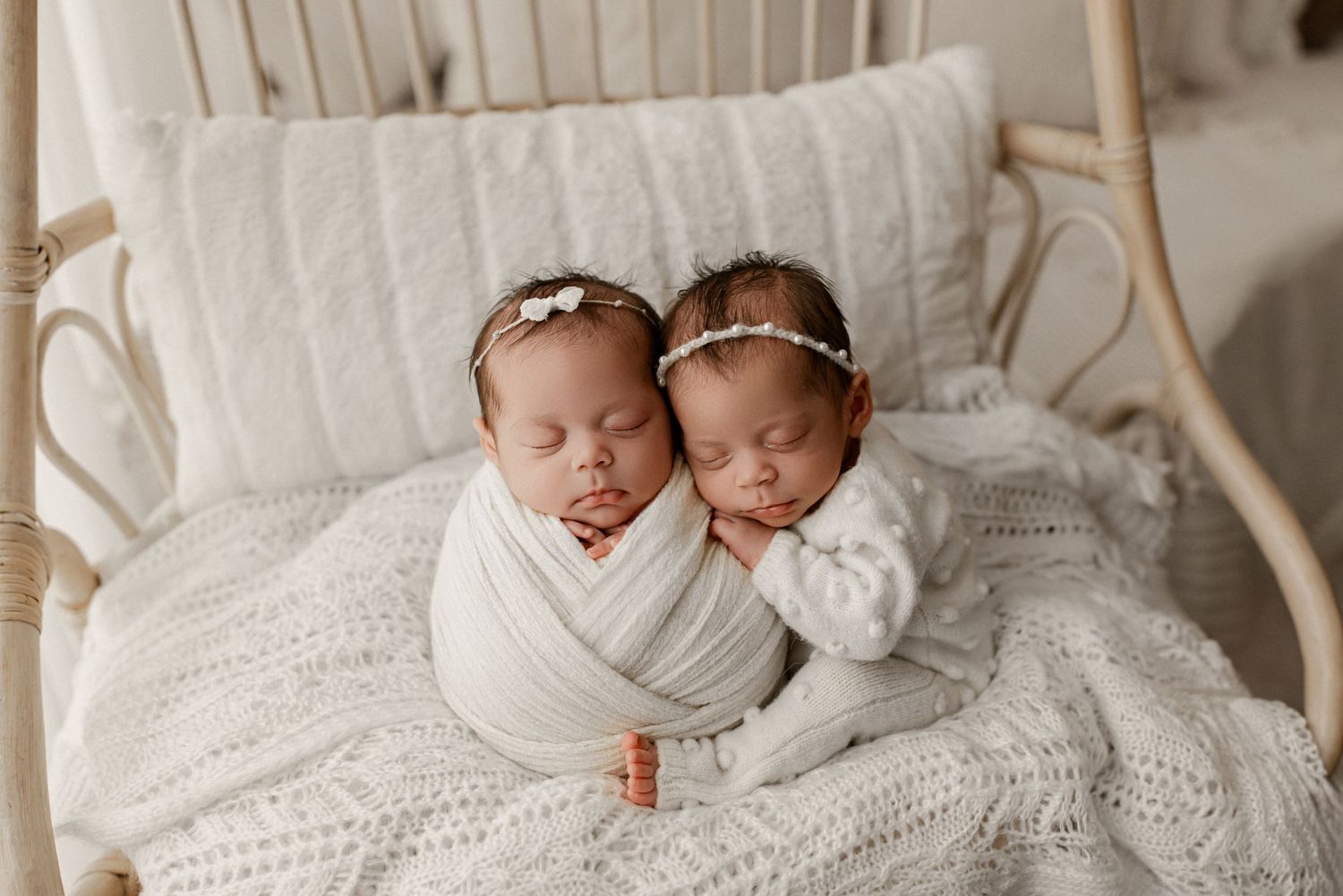Two sleeping newborn babies swaddled in white lying together on a white knit blanket in a rattan chair.