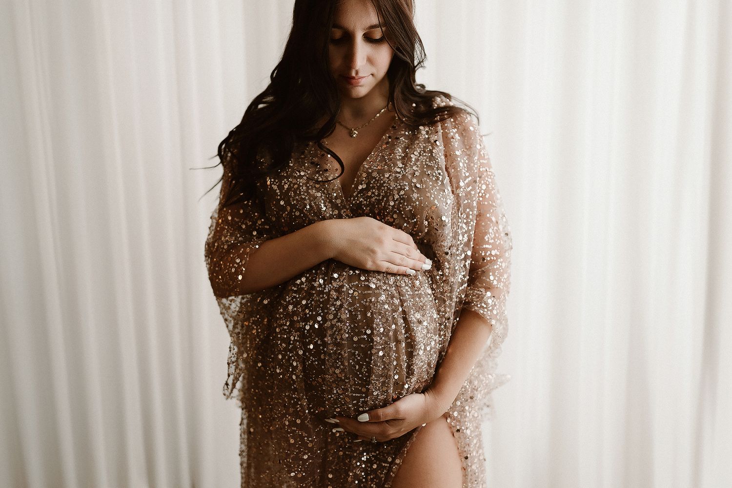 Silhouetted pregnant figure in a sparkly sequined dress stands elegantly against white curtain backdrop.