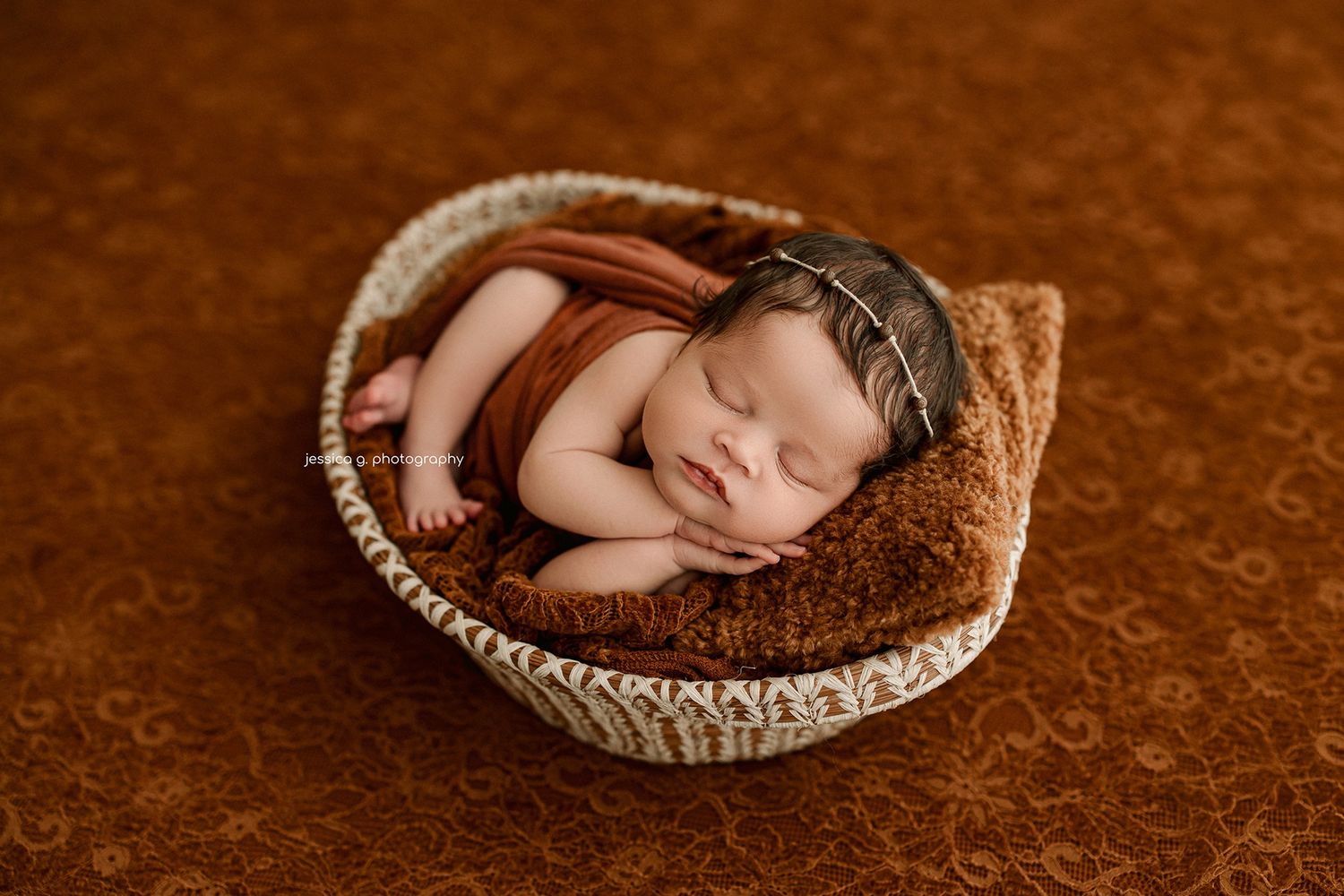 Sleeping newborn baby in brown wrap and aviator hat posed in woven basket on warm brown floral background.