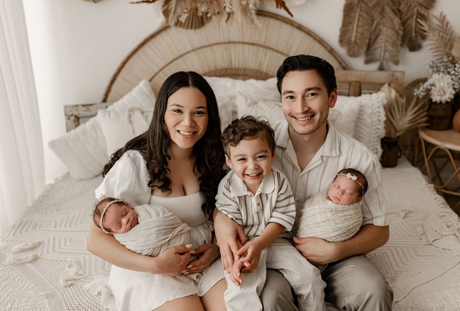 A family sitting together on a white bed with neutral decor, holding two newborn babies.