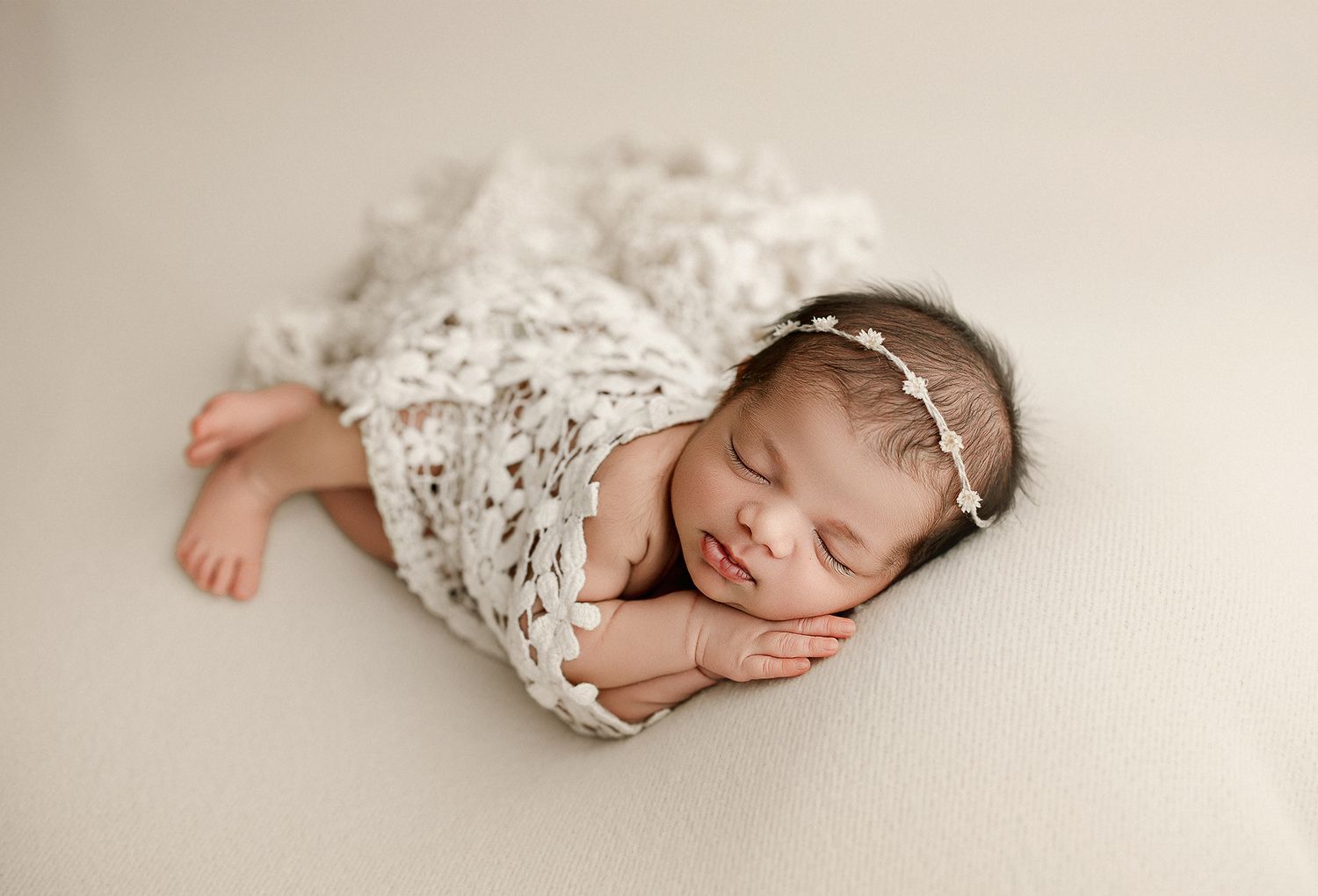Sleeping newborn baby girl wrapped in white lace swaddle with floral headband on cream background.