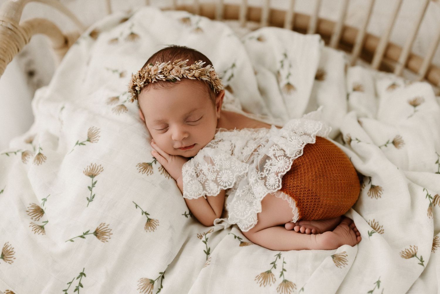 Sleeping newborn wearing delicate lace dress rests on white floral blanket with dainty flower crown.