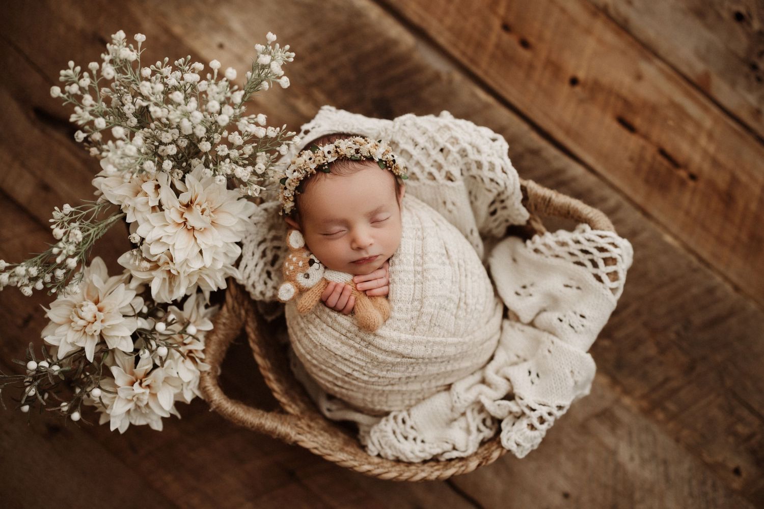 A sleeping newborn wrapped in cream knit fabric rests in a rustic basket with delicate white flowers on wooden floor.