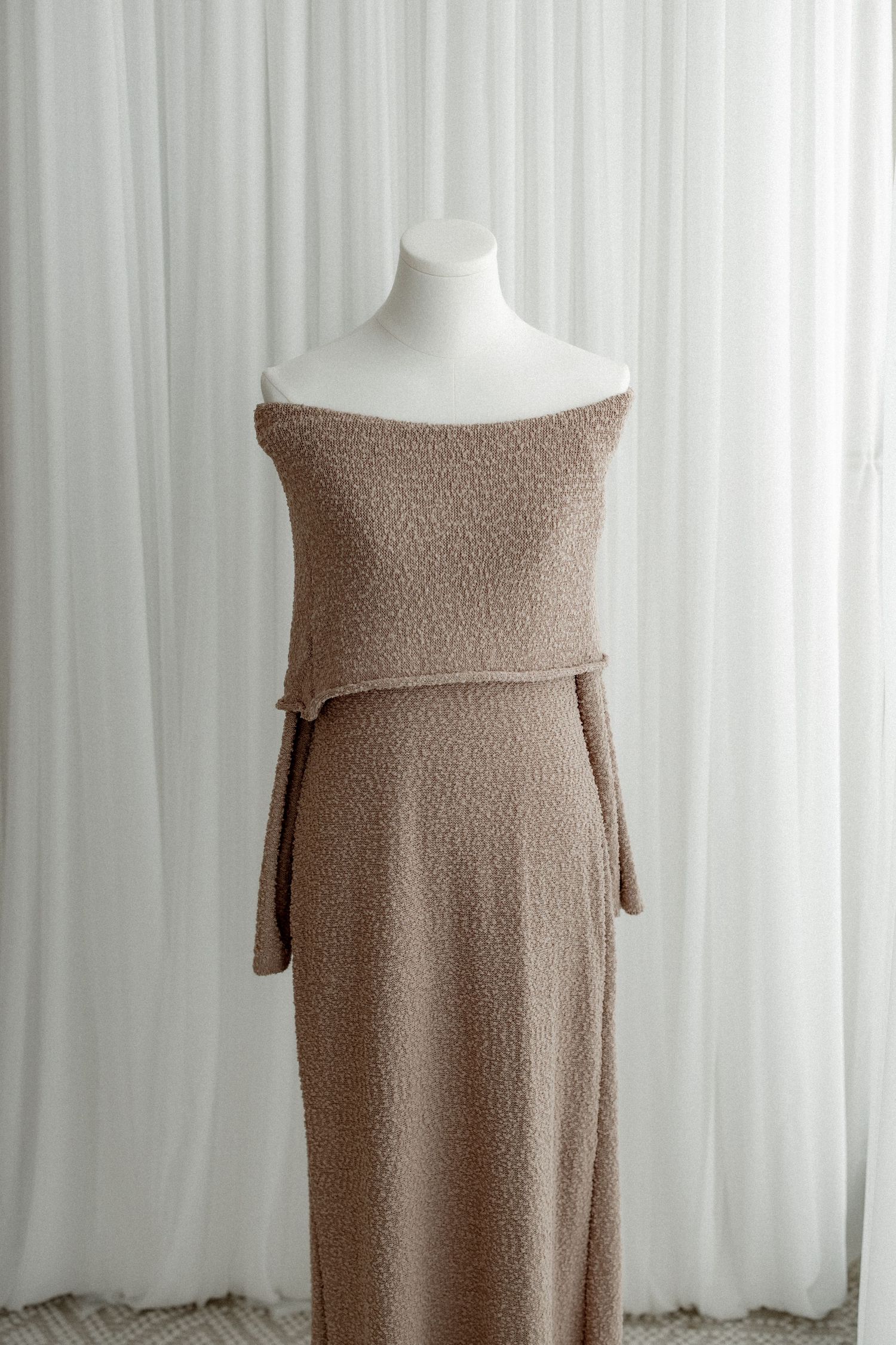 Brown knit off-shoulder dress displayed on white mannequin against white curtain background.