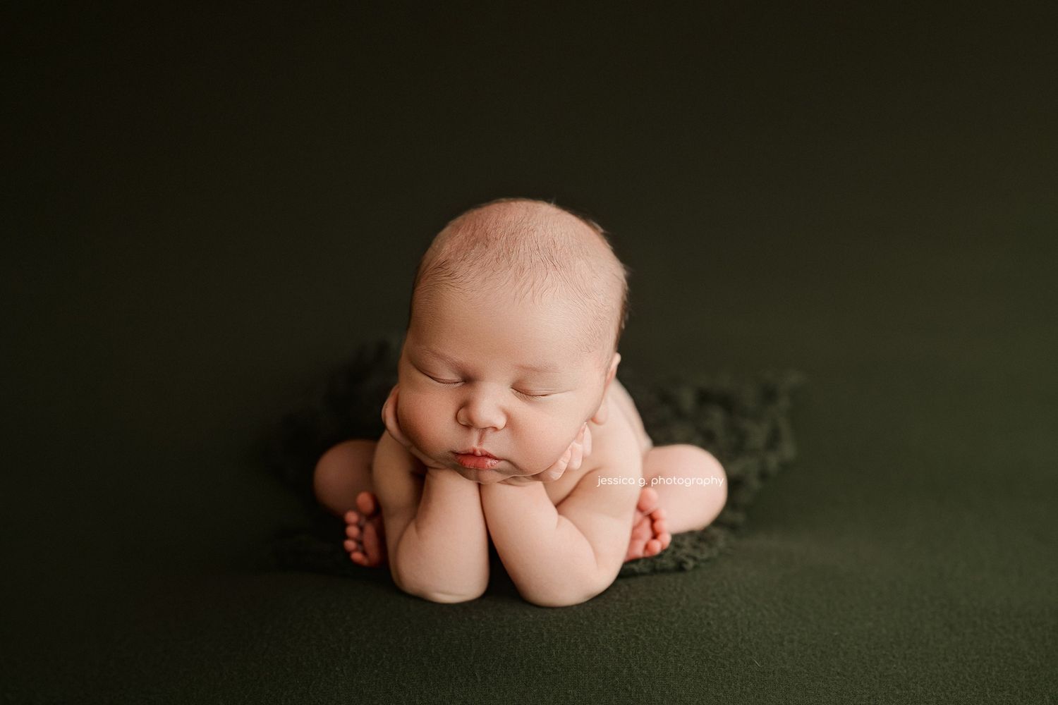 Sleeping newborn baby posed on dark green backdrop in froggy position during professional photo session.