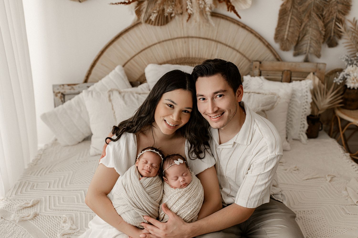 A family rests together on a white bed with twin newborns wrapped in neutral swaddles.