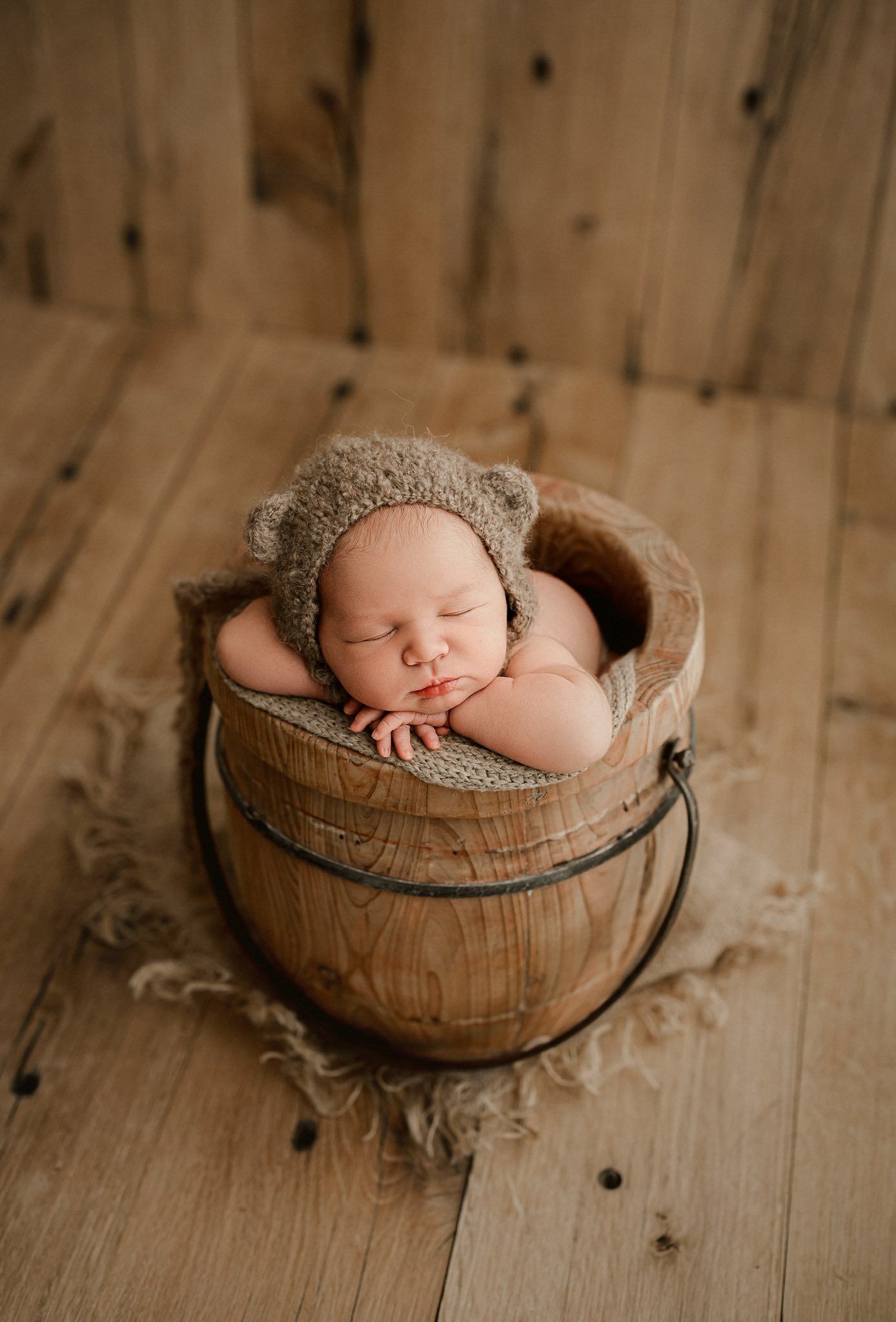 A sleeping newborn rests peacefully in a rustic wooden bowl lined with soft fabric on a wooden floor.