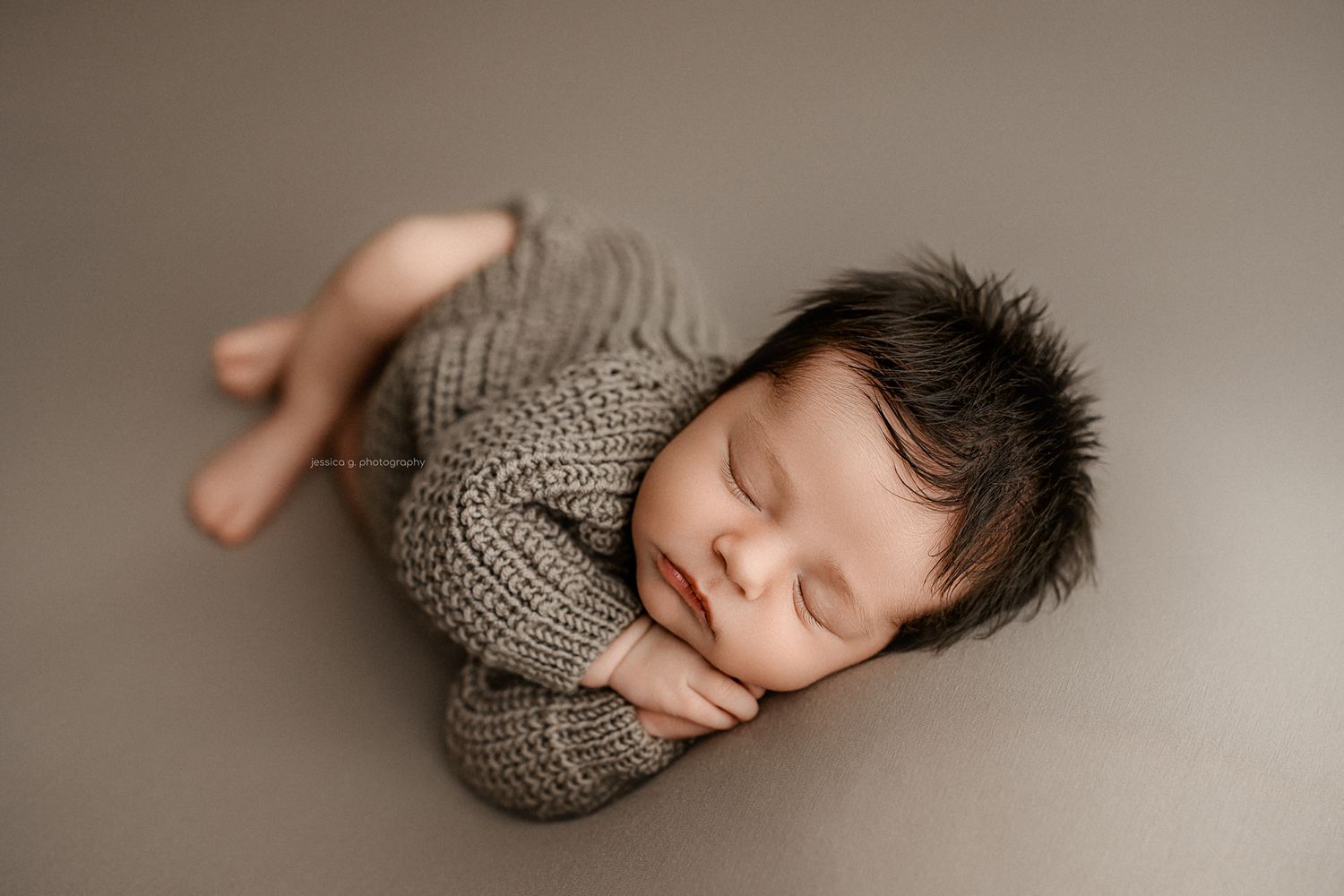 Newborn baby sleeping peacefully in gray knit wrap on brown backdrop.